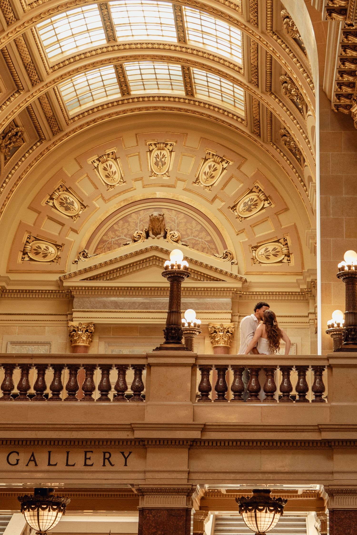 Couple sharing a romantic moment on a balcony inside an ornate gallery with golden arched ceiling.