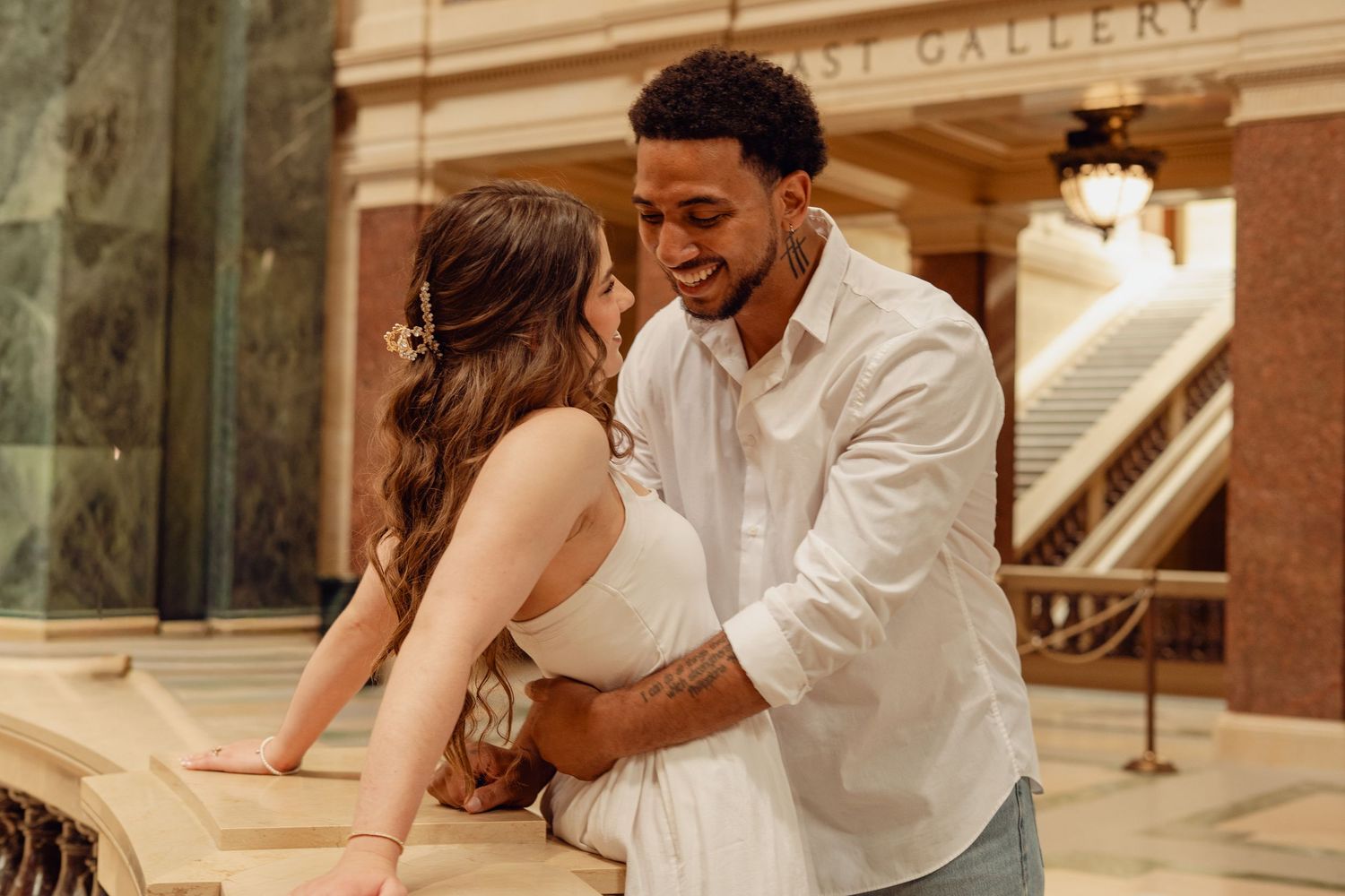 A couple shares a romantic embrace inside an elegant gallery building with marble architecture.