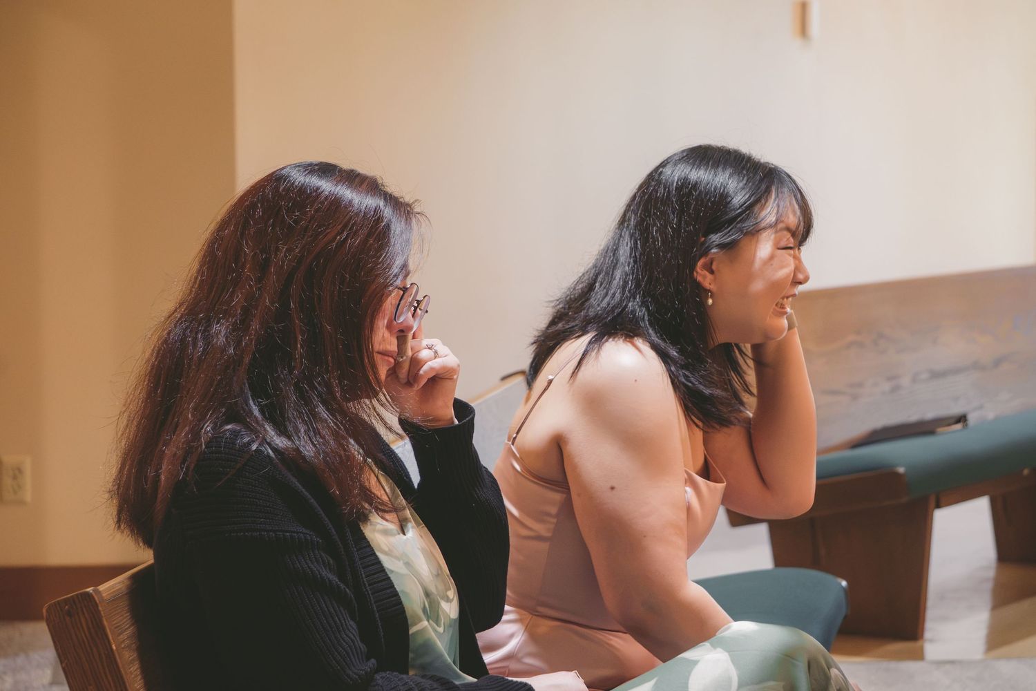 Two young Asian women sitting side by side, laughing and covering their mouths in an elegant indoor setting.