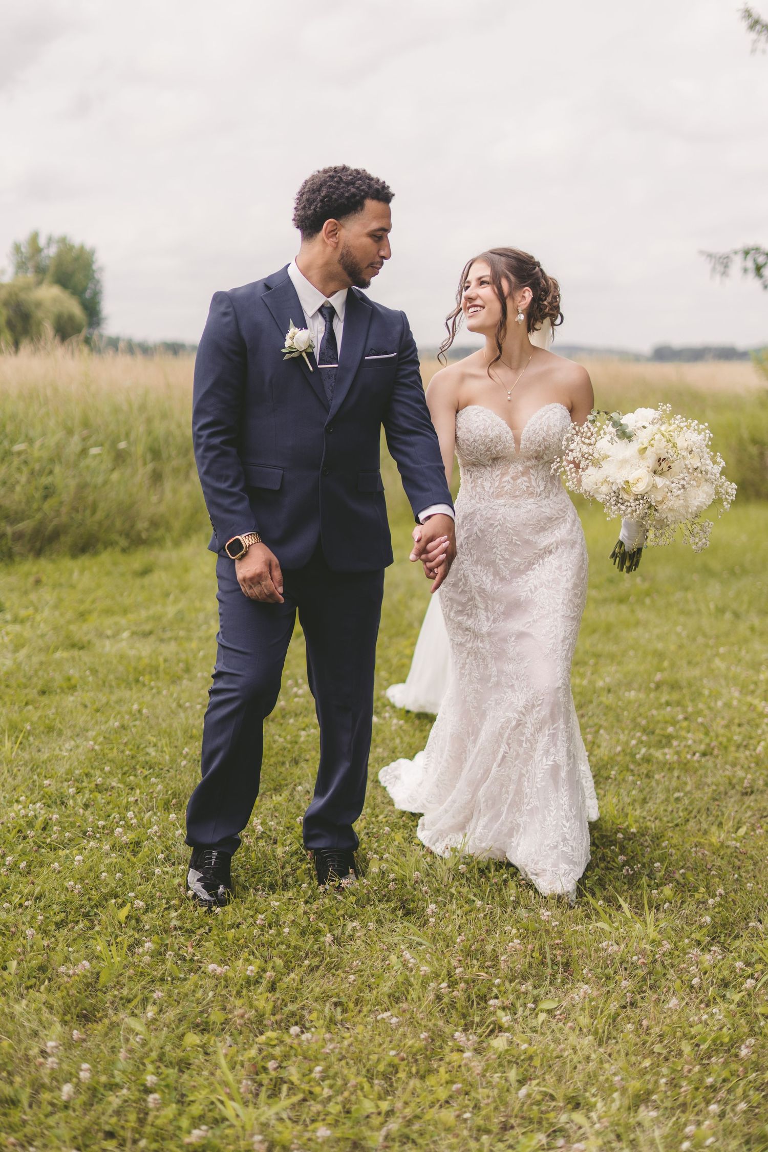 A bride and groom walk hand-in-hand through a green meadow, she in a lace gown, he in a navy suit.