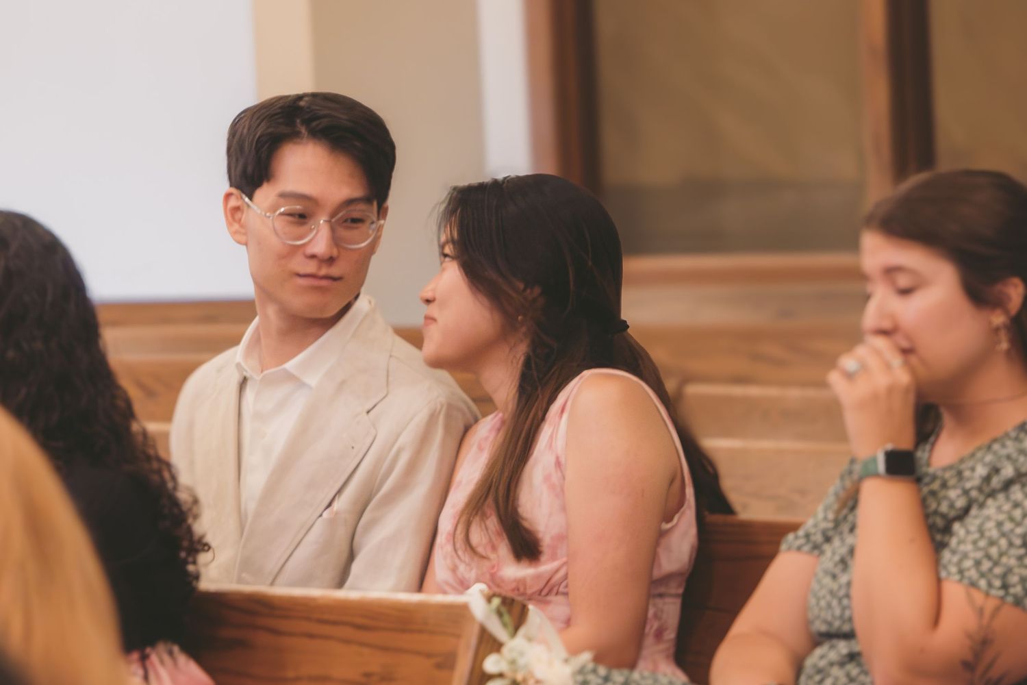 Guests in formal attire sit in church pews during a wedding ceremony, with a man in beige suit and woman in pink dress.