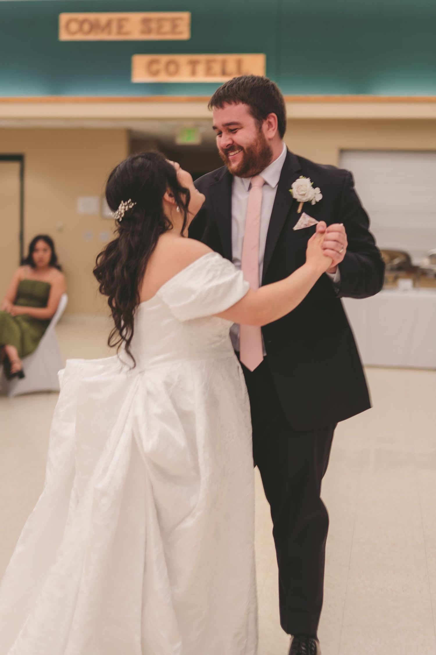 Bride and groom share their first dance, groom in dark suit with boutonniere, bride in white gown.