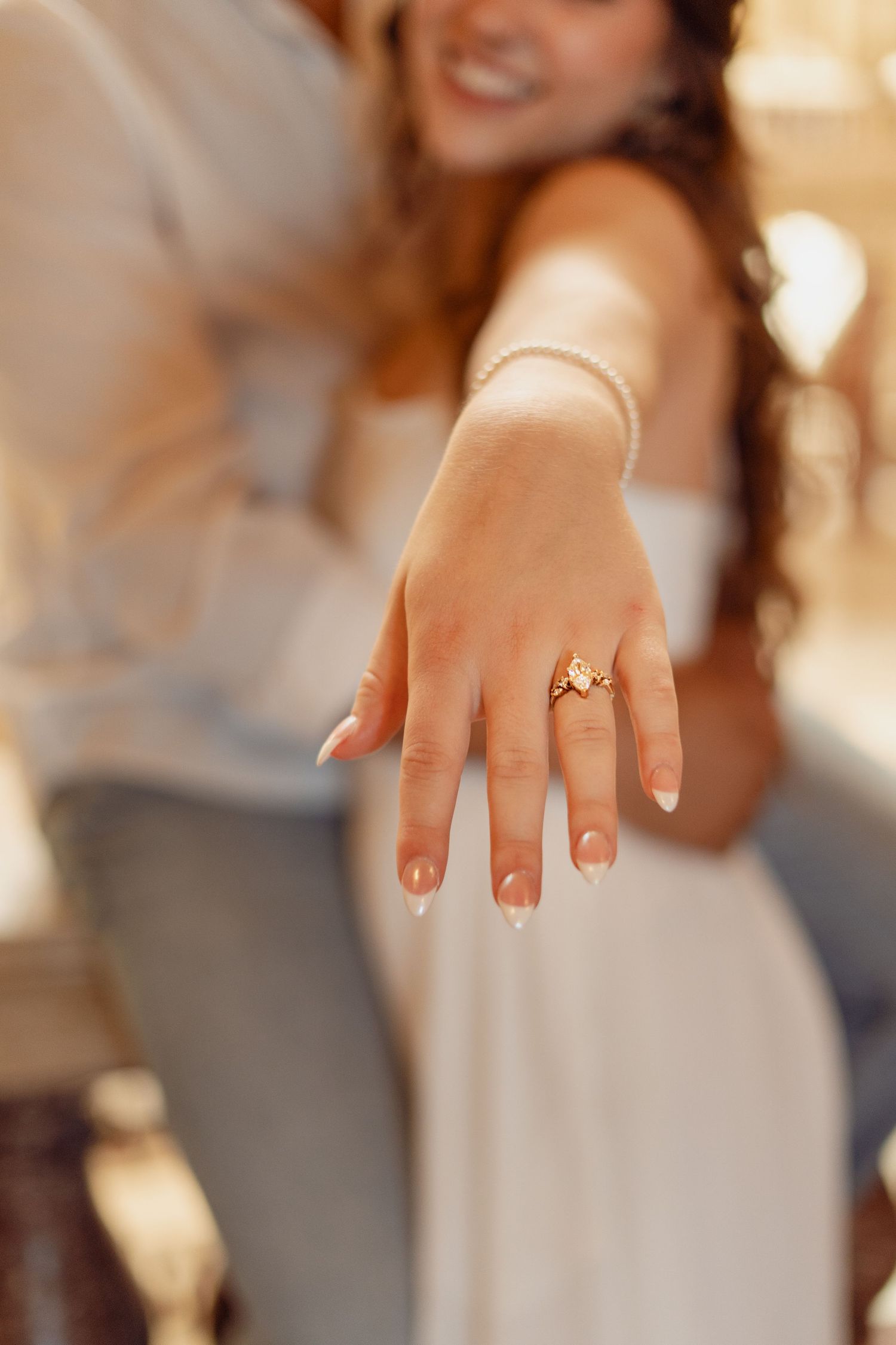 Close-up of bride showing gold engagement ring with French manicure, pearl bracelet visible in background.