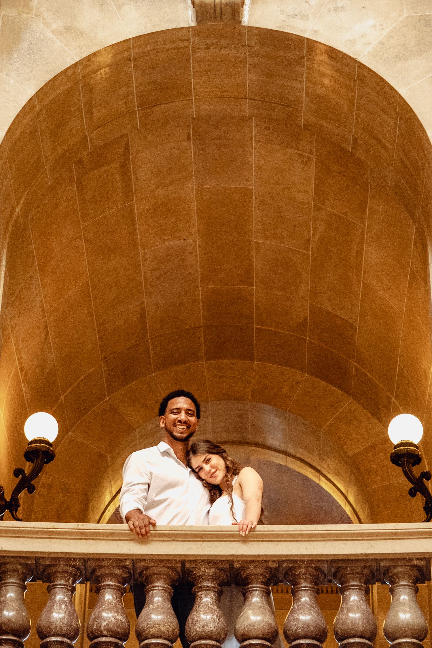 Romantic interracial couple embracing on ornate balcony under golden arched ceiling with vintage wall sconces.