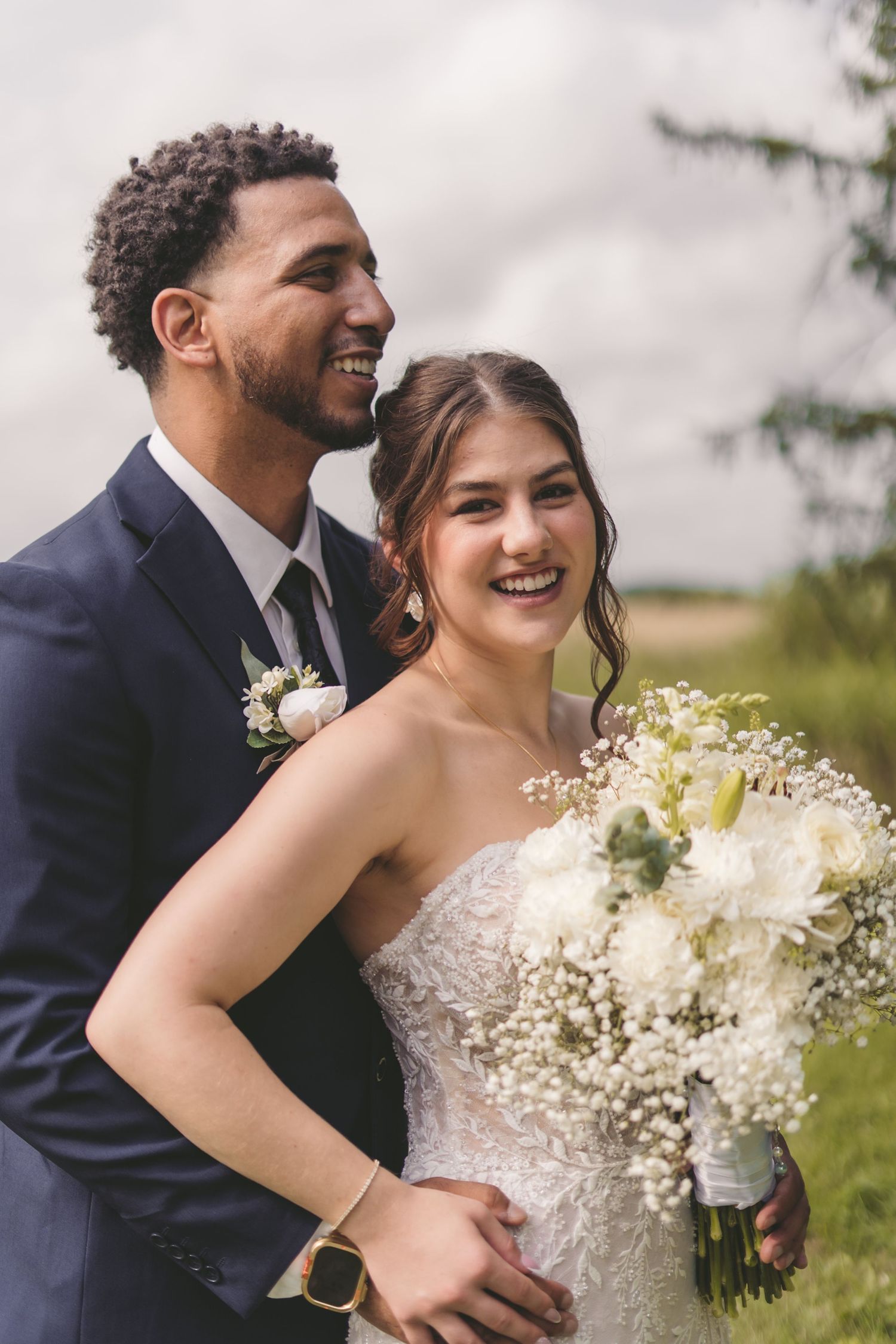 A smiling bride holding white flowers stands with a groom in a navy suit outdoors on their wedding day.