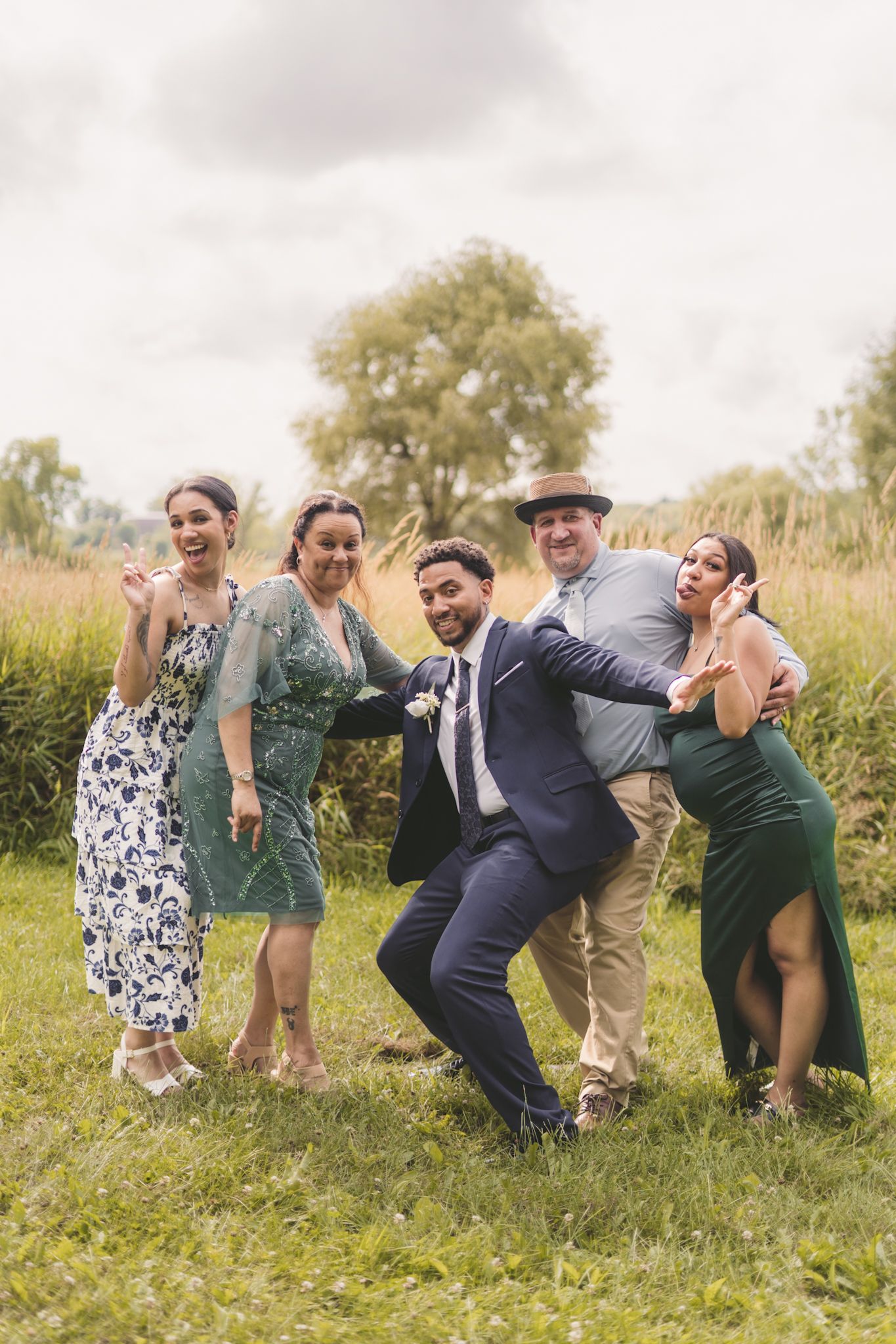 A group of five joyful wedding guests pose and laugh together on a green lawn with tall grass and trees behind them.