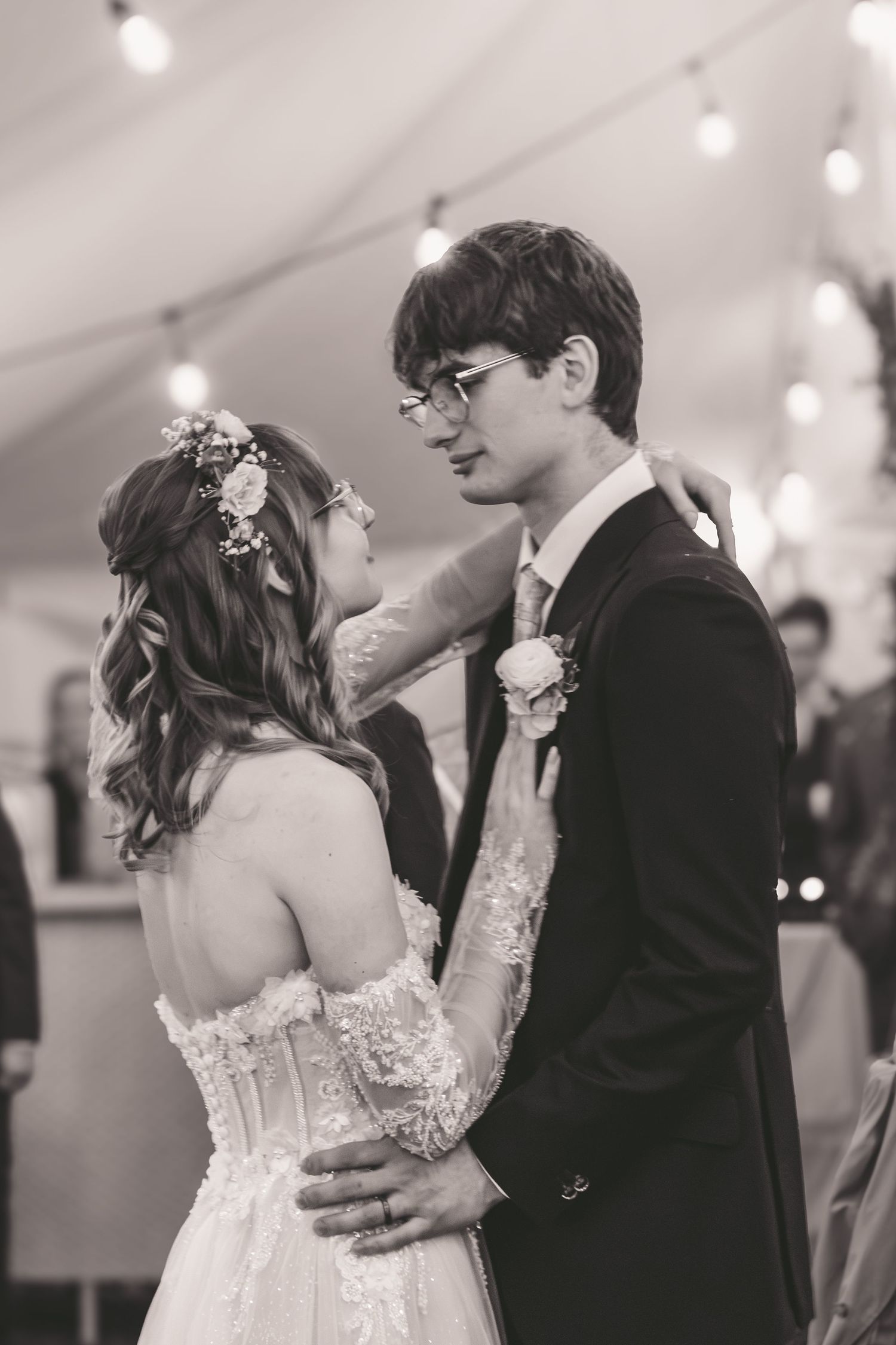 Black and white photo of bride and groom sharing their first dance under string lights at wedding reception.