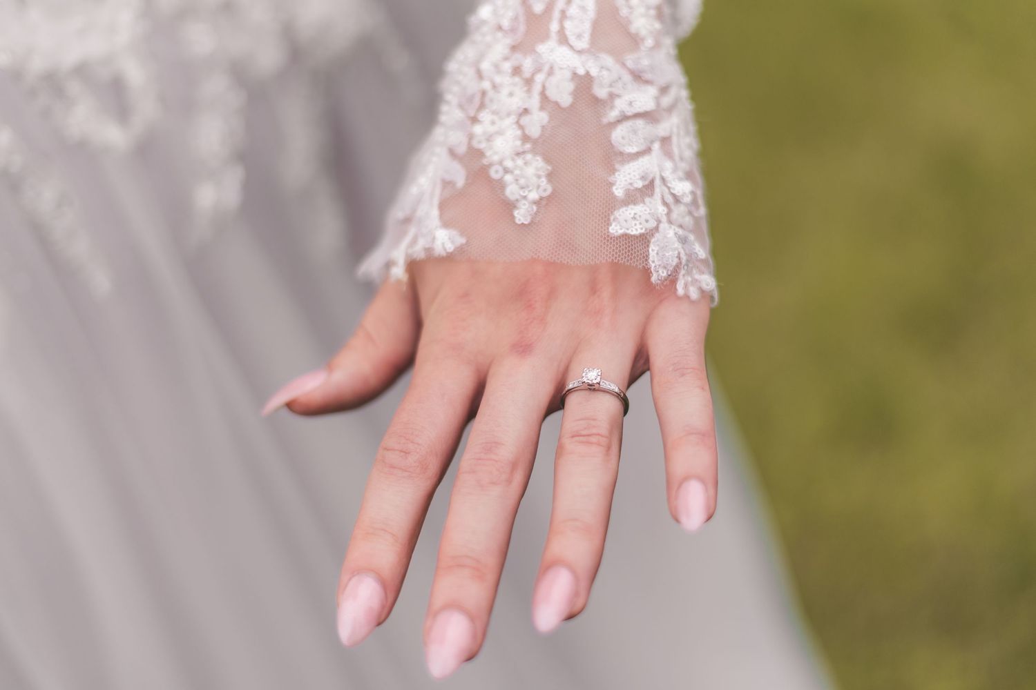Bride showcasing elegant diamond engagement ring on manicured hand with pink nails, wearing lace wedding dress sleeve.
