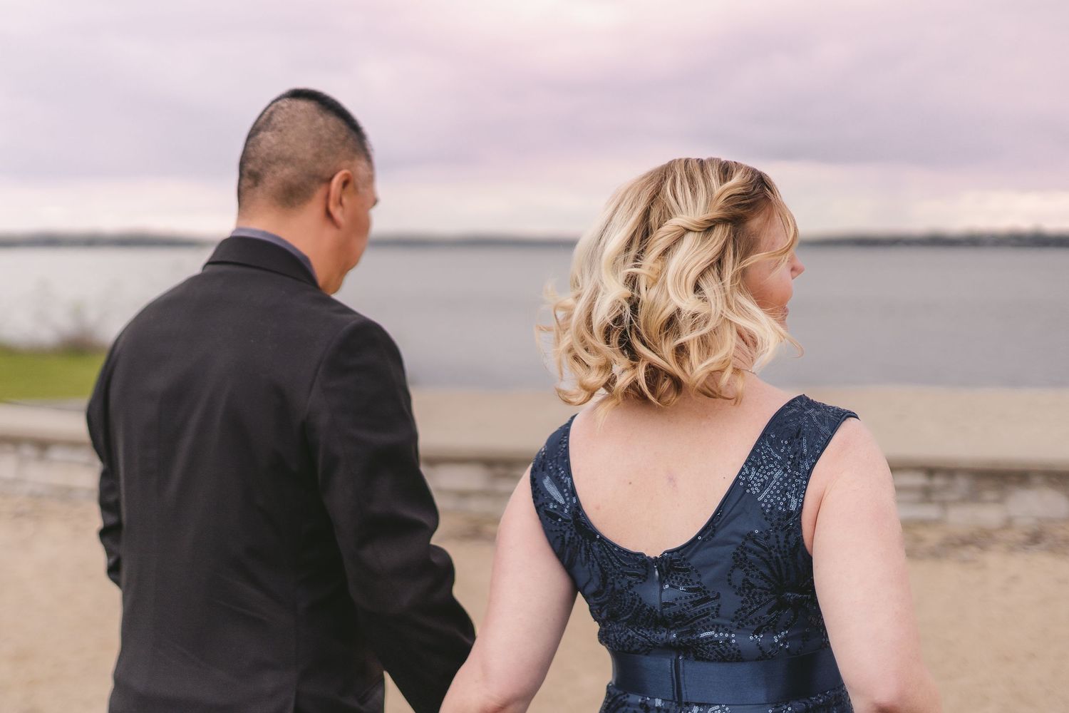 Couple holding hands in formal wear by waterfront at dusk, woman in navy sequin dress, man in black suit.