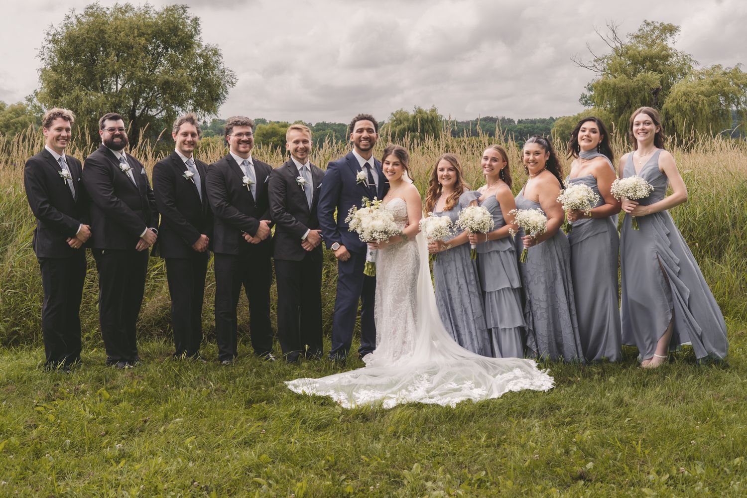 Wedding party posing outdoors on grass, bride in white gown with groomsmen in black suits and bridesmaids in blue dresses.