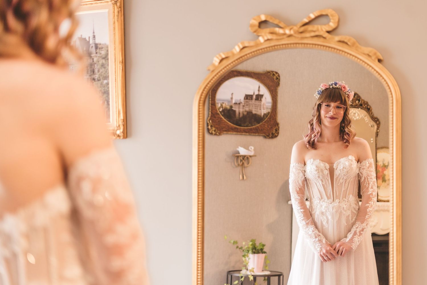 Bride in lace wedding gown admiring her reflection in an ornate gold mirror.