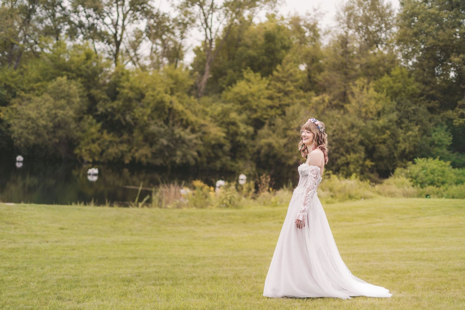 Bride in elegant white off-shoulder gown standing gracefully on green lawn with trees and pond in background.