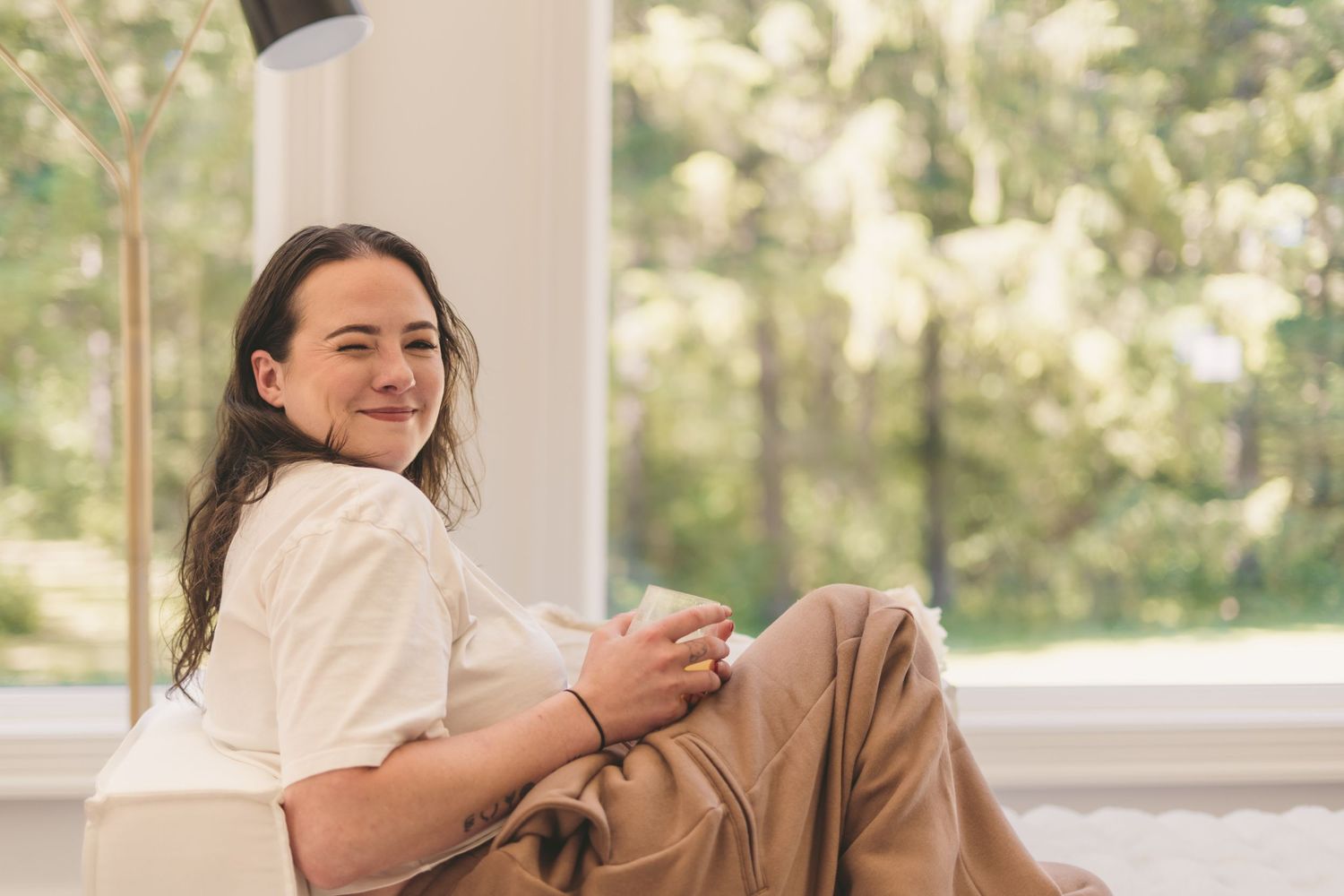 Young woman with long hair smiling while relaxing in a chair, holding a mug near a bright window with trees.