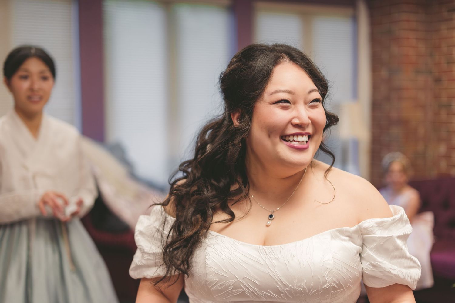 Smiling Asian bride in white off-shoulder gown laughing joyfully at wedding reception.