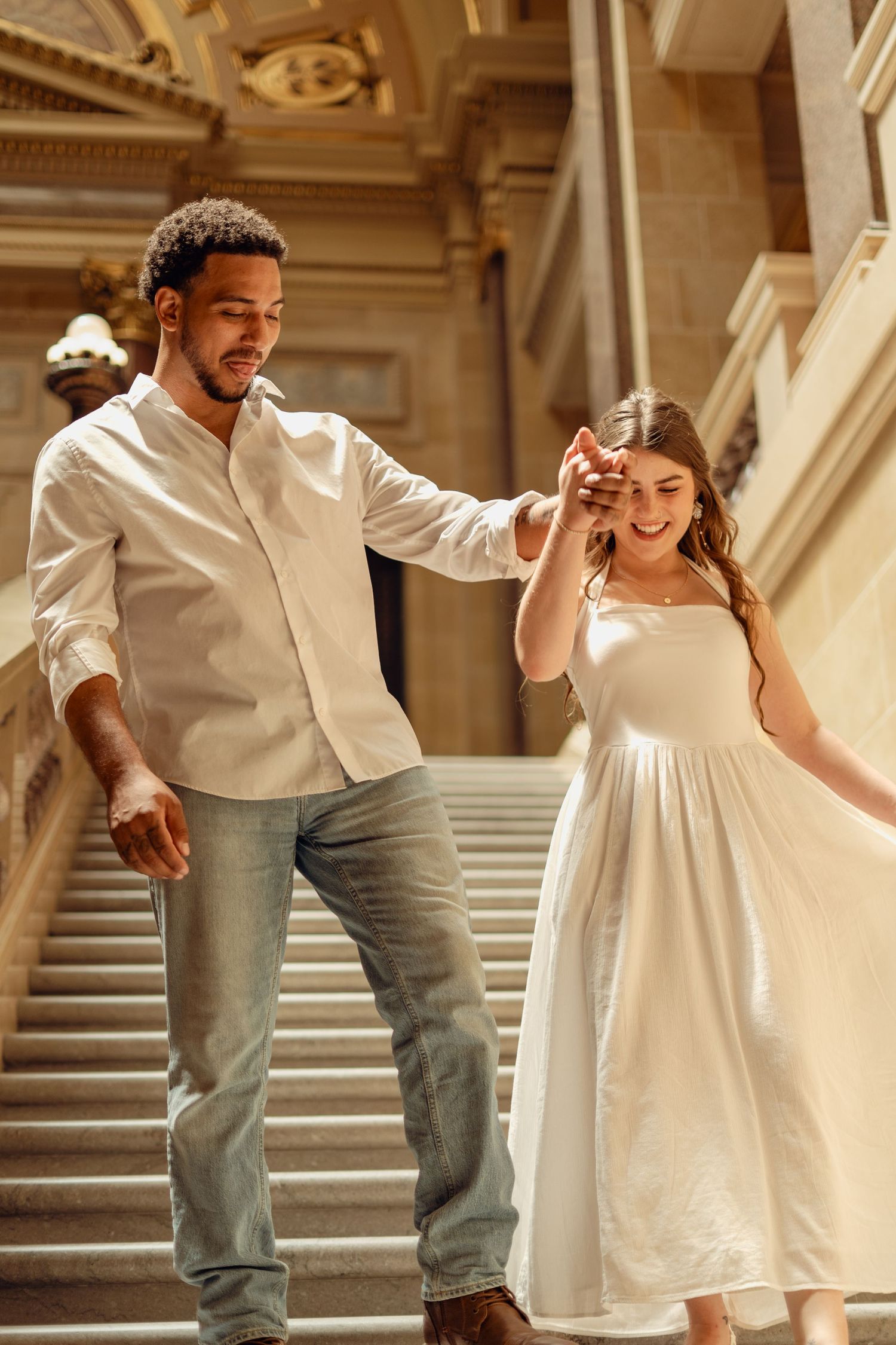 Interracial couple holding hands walking down grand staircase, man in white shirt and jeans, woman in white sundress.