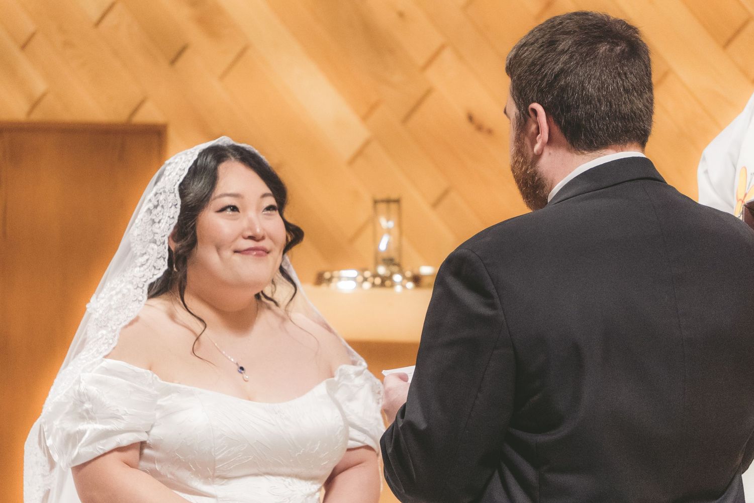 Bride in white lace gown and veil smiling at groom during wedding ceremony in elegant wood-paneled chapel.