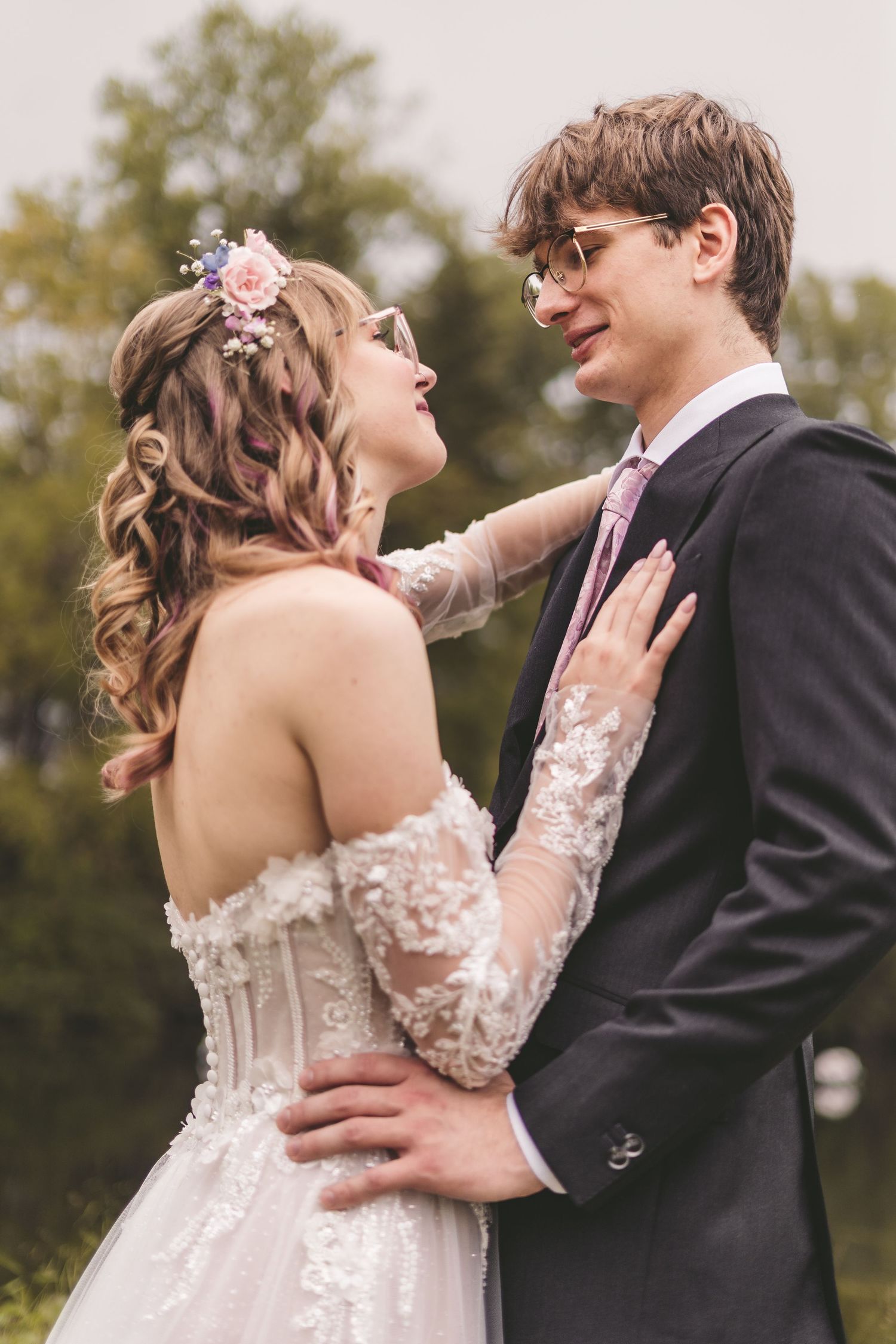 Bride in lace corset gown with floral crown laughing with groom in dark suit outdoors.