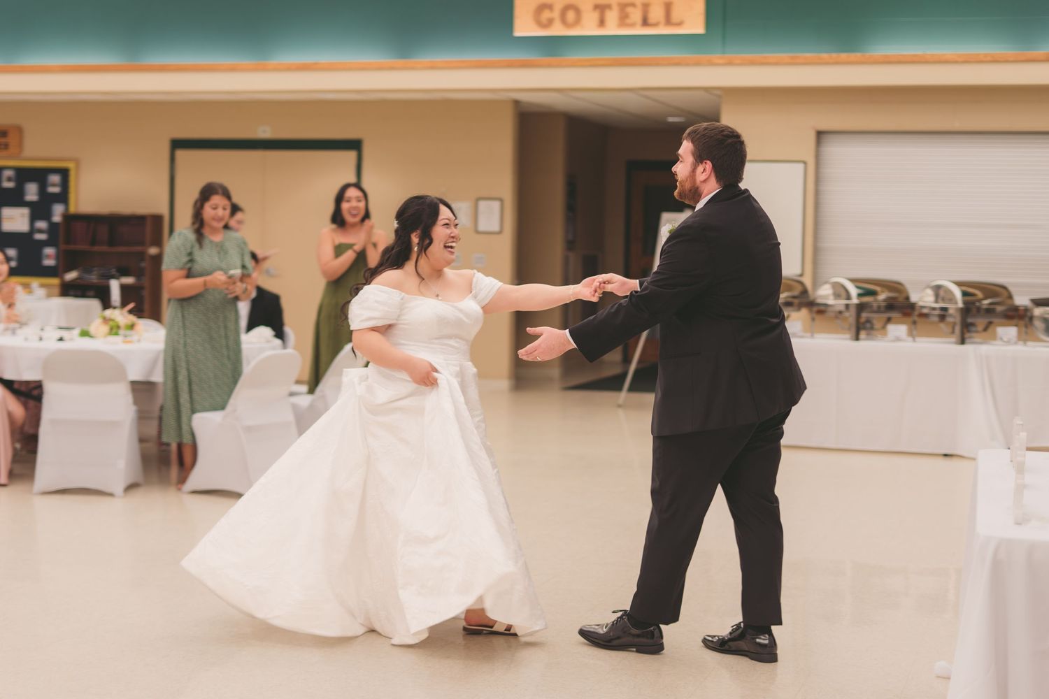 Bride and groom share their first dance at wedding reception while guests in green dresses look on and applaud.
