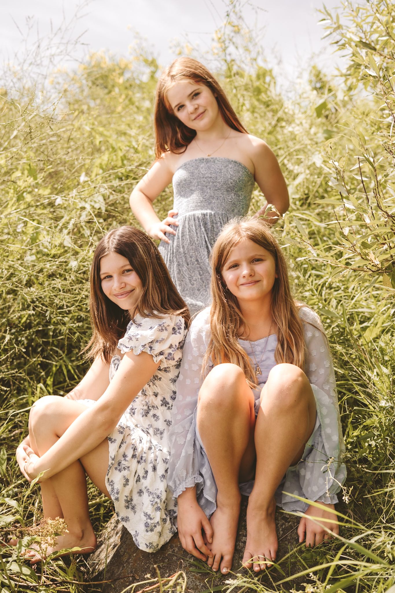 Three girls in summer dresses pose outdoors among tall green plants on a sunny day.