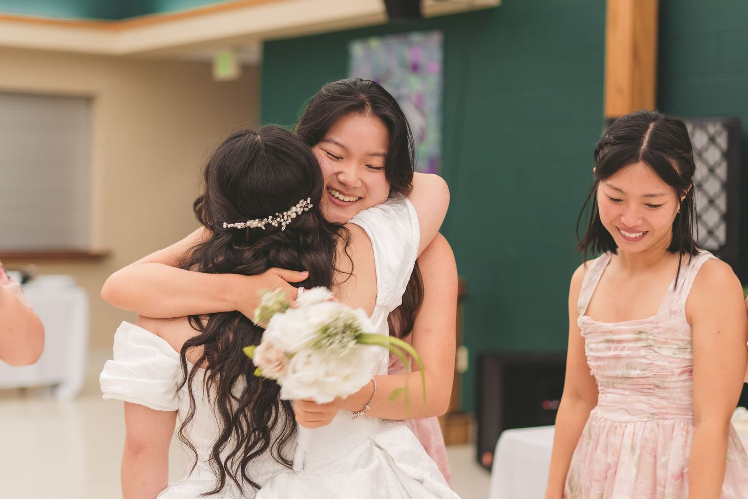 Bride receives a warm hug from a guest while holding a white bouquet at a wedding reception.