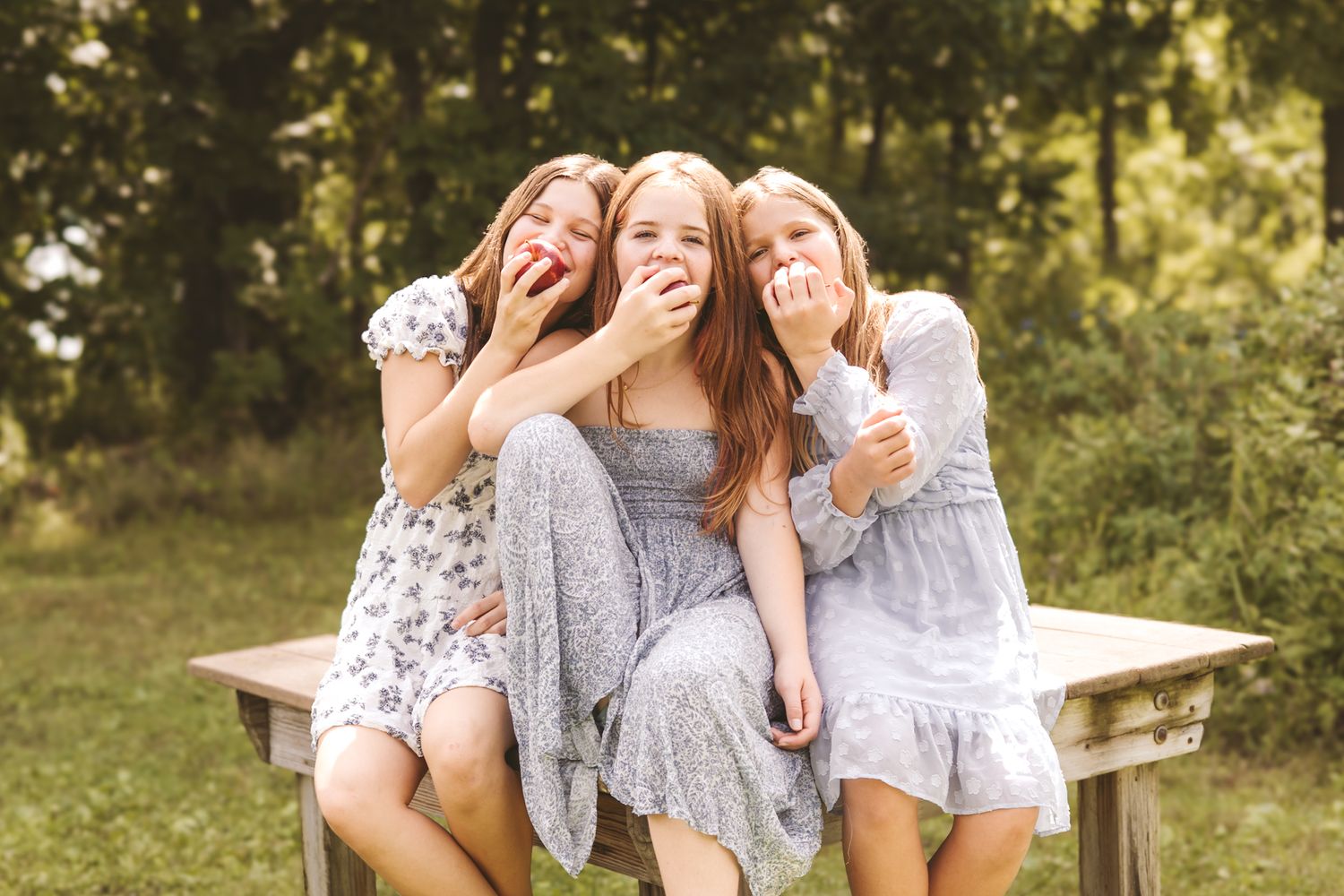 Three girls sit outdoors laughing and eating apples together on a rustic wooden bench.