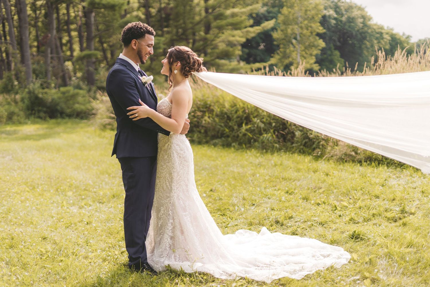Bride and groom embrace outdoors on green grass, her lace wedding dress veil flowing in the wind.