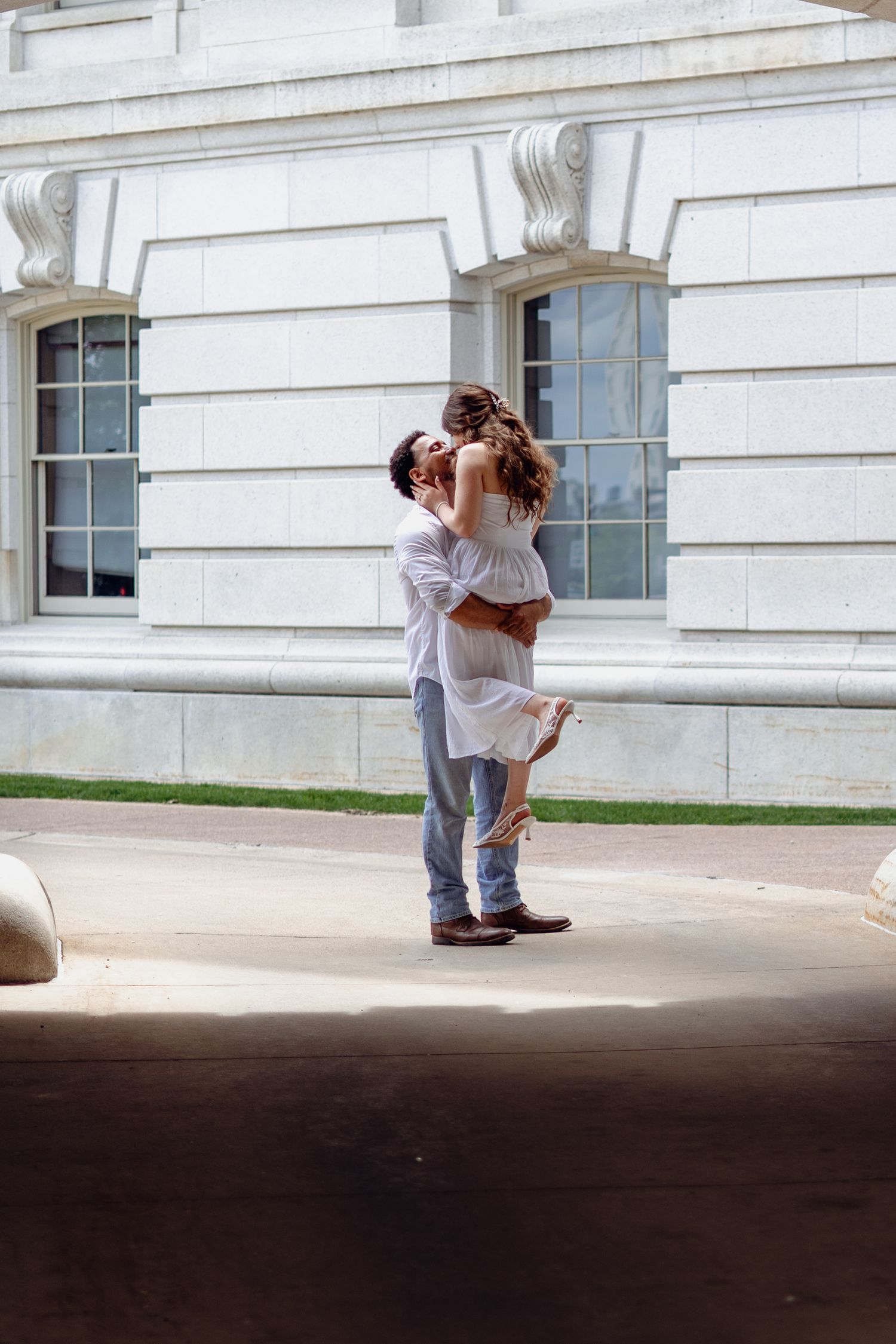Romantic couple sharing a kiss as man lifts woman in front of a white neoclassical building outdoors.