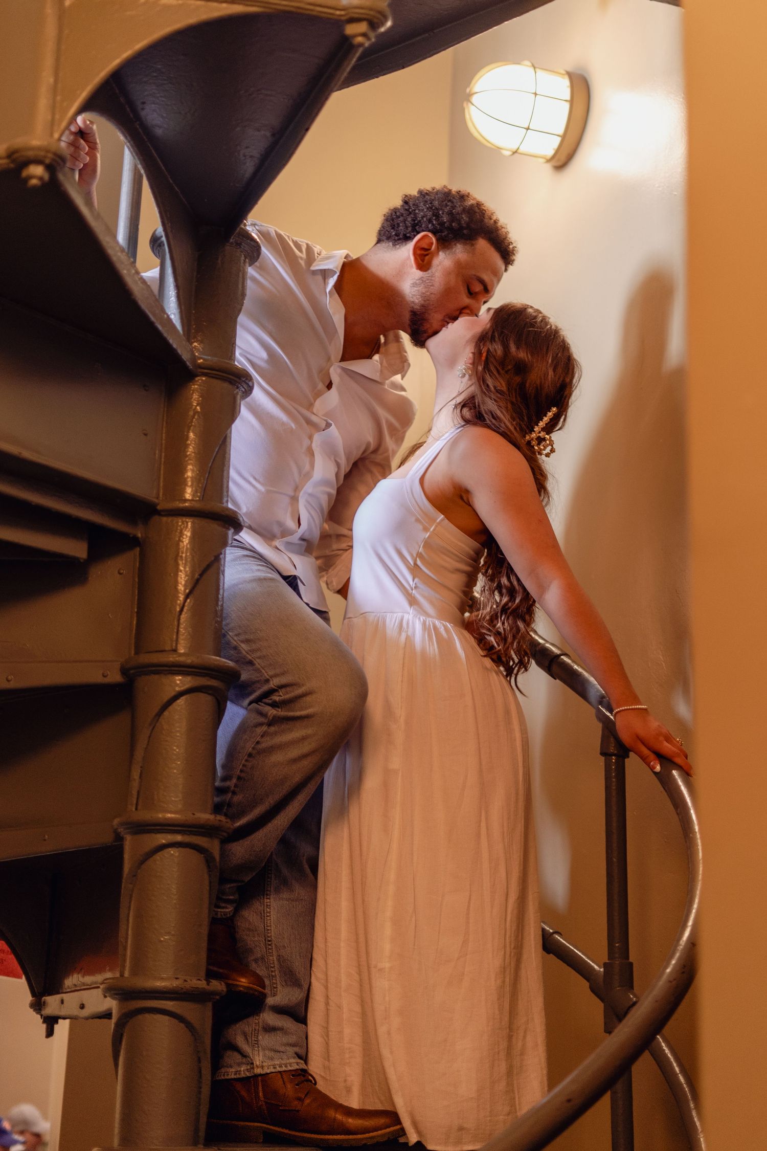 Romantic interracial couple kissing on a spiral staircase in warm ambient lighting.