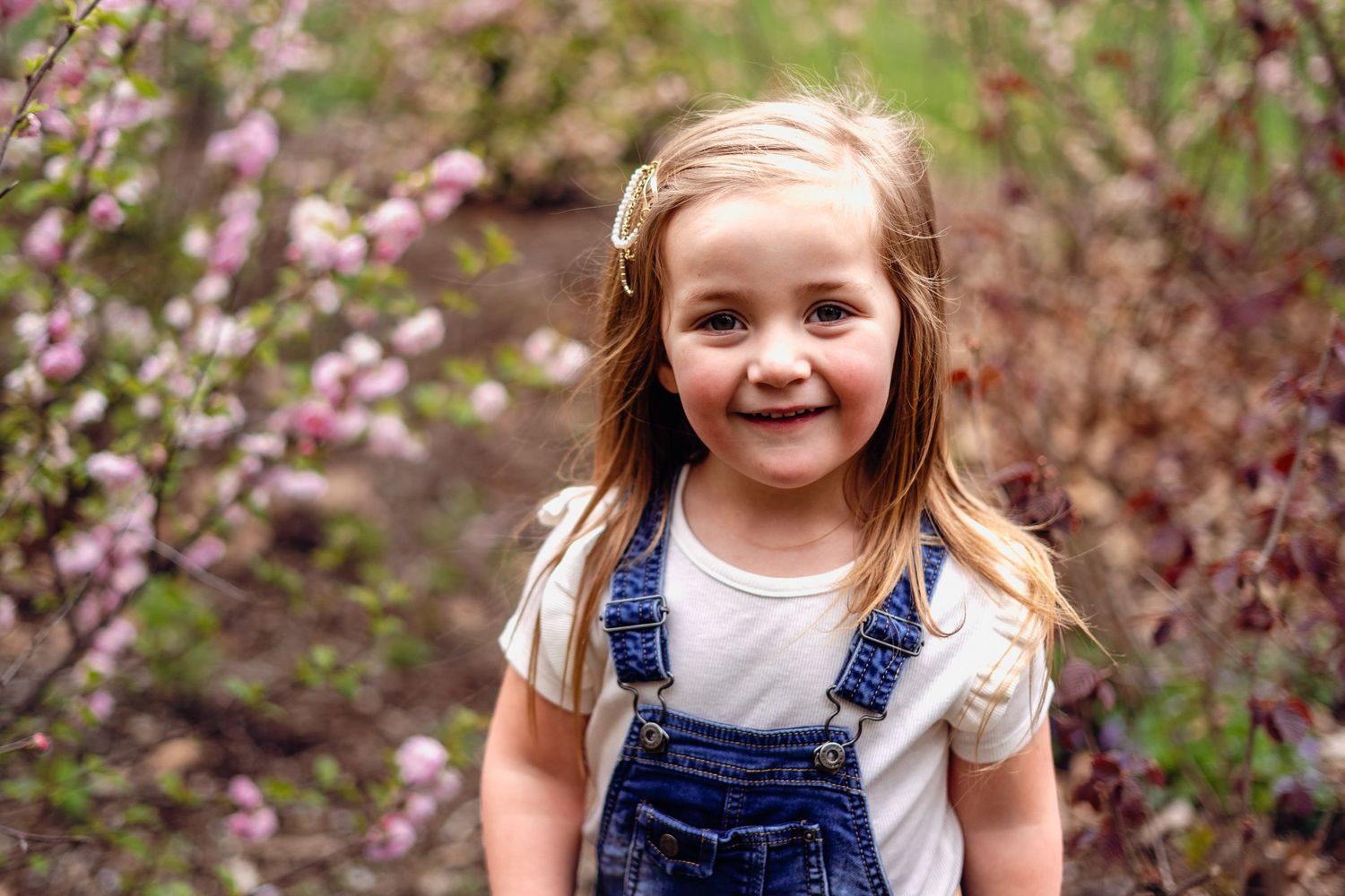 Child in denim overalls smiling among purple spring flowers.