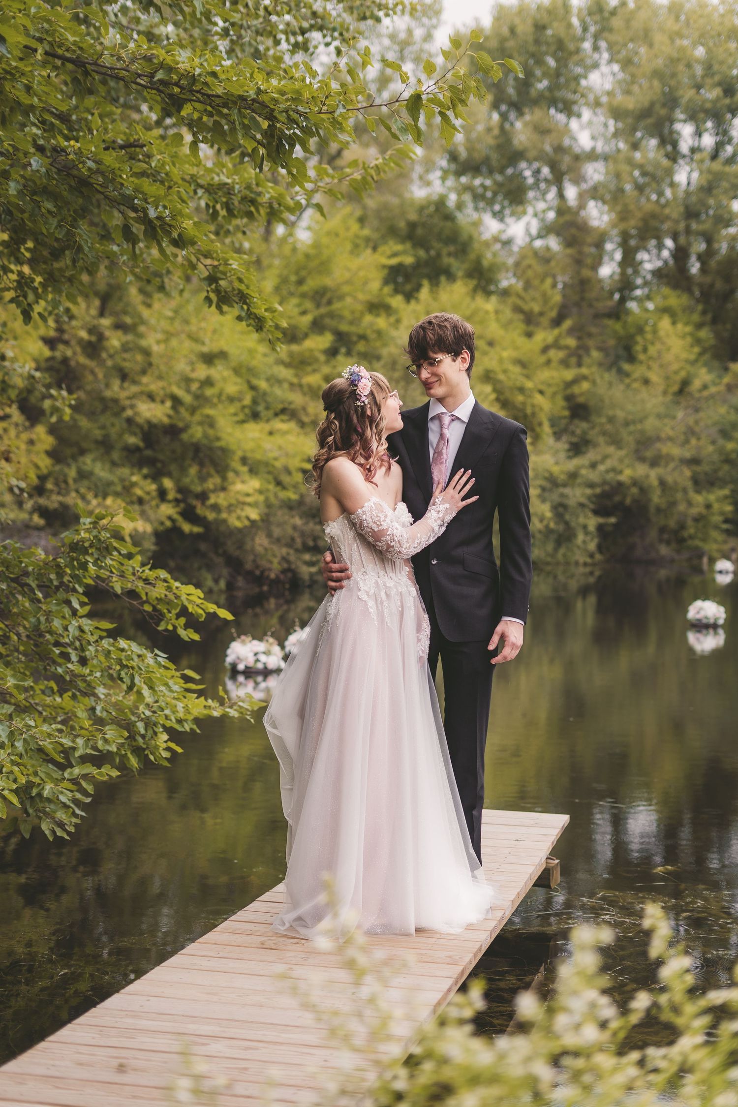 Bride and groom sharing romantic moment on wooden dock surrounded by lush green trees and lake.