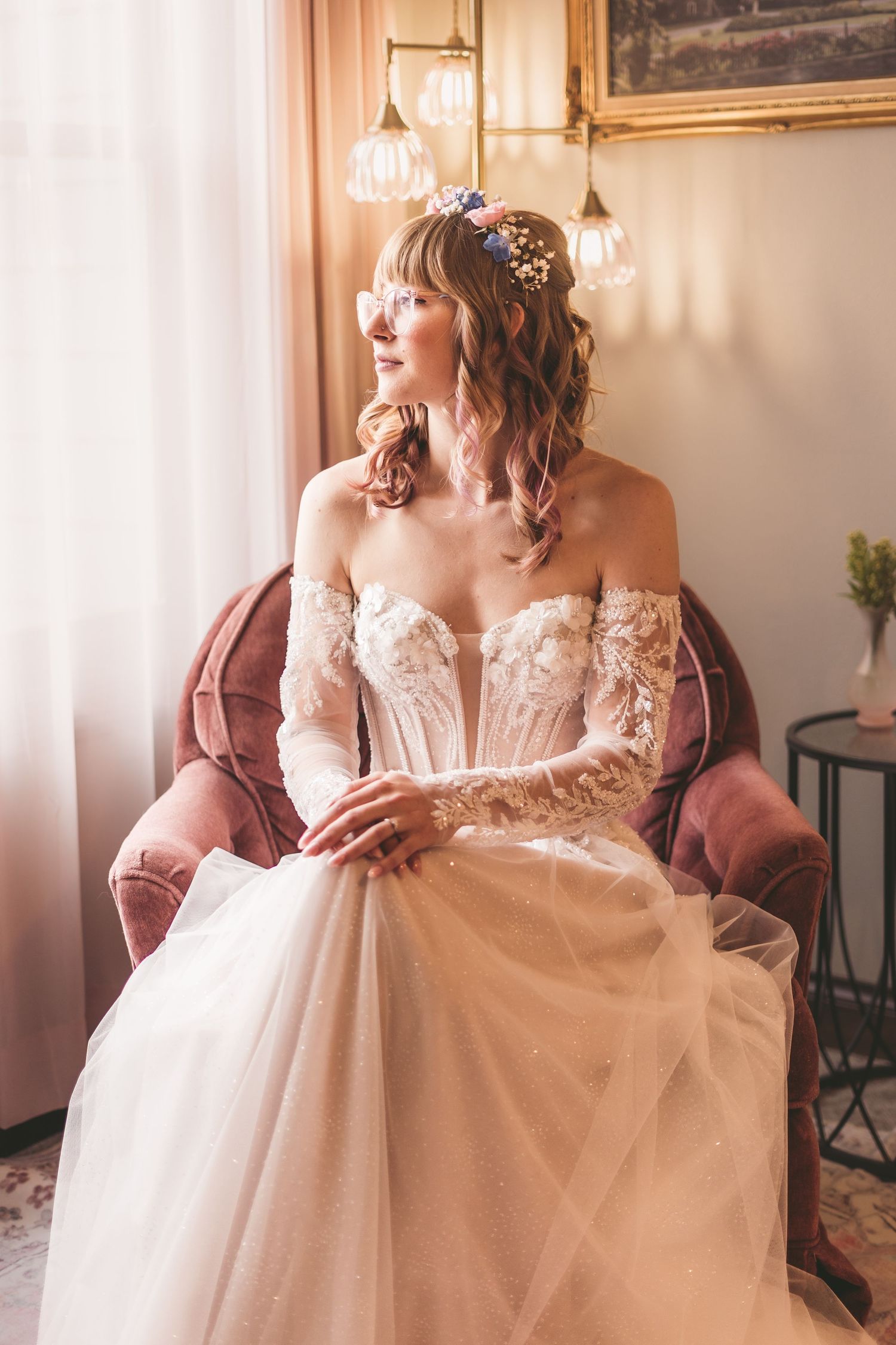 Bride in lace off-shoulder wedding gown with floral crown sits in pink velvet chair by sunlit window.