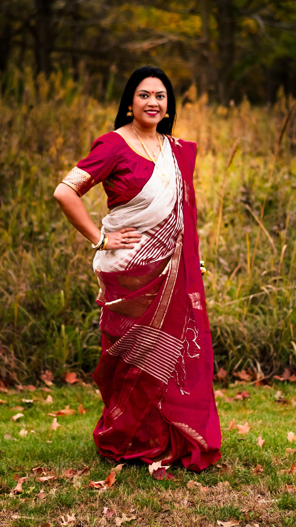 Woman in elegant red and white saree with gold accents poses gracefully outdoors in a lush green setting.