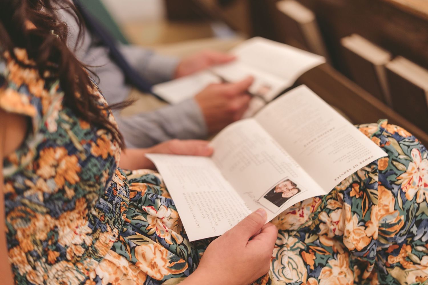 Woman in floral dress reading a wedding program booklet while seated in a church pew during a ceremony.
