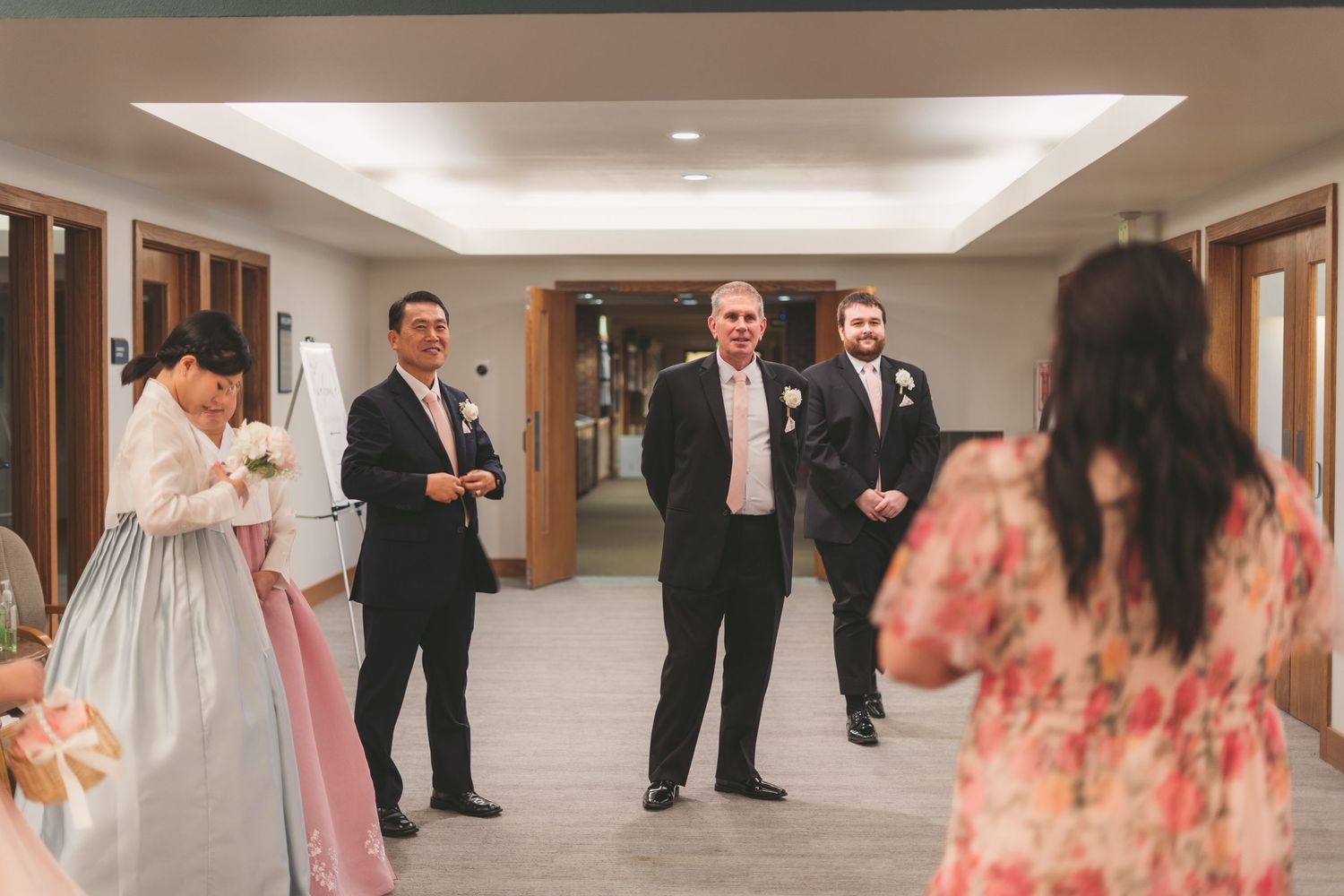 Wedding guests in formal attire gather in a hallway, with a woman in traditional Korean dress standing nearby.