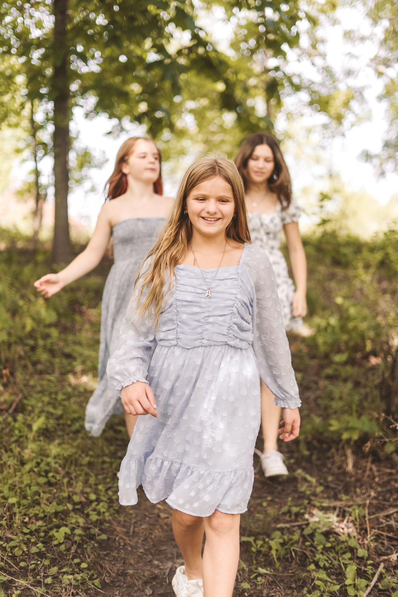 Three girls in blue dresses walking through a sunlit forest, smiling and enjoying a peaceful outdoor stroll.
