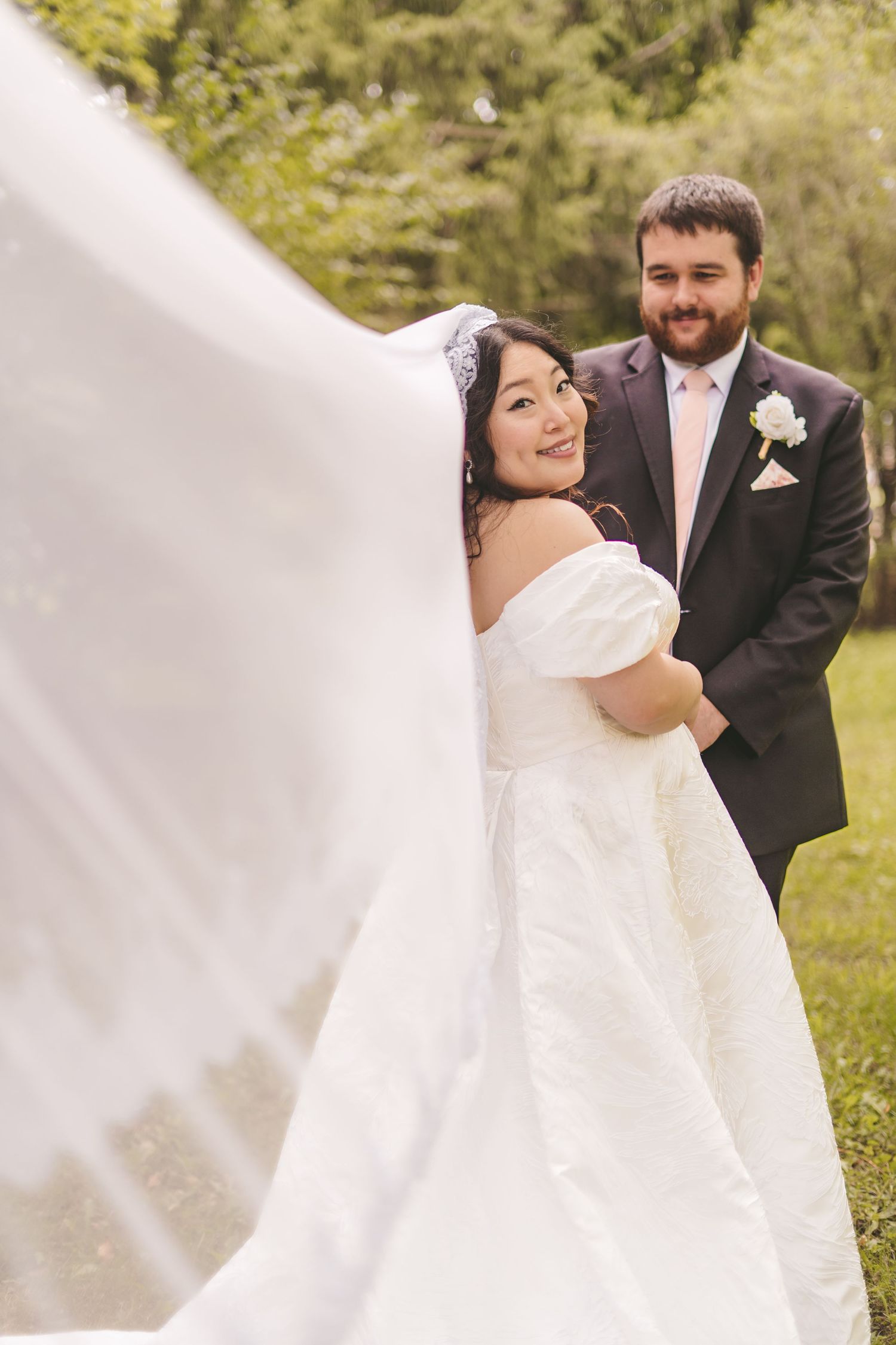 Bride in white off-shoulder gown with flowing veil poses with groom in dark suit and pink tie outdoors.