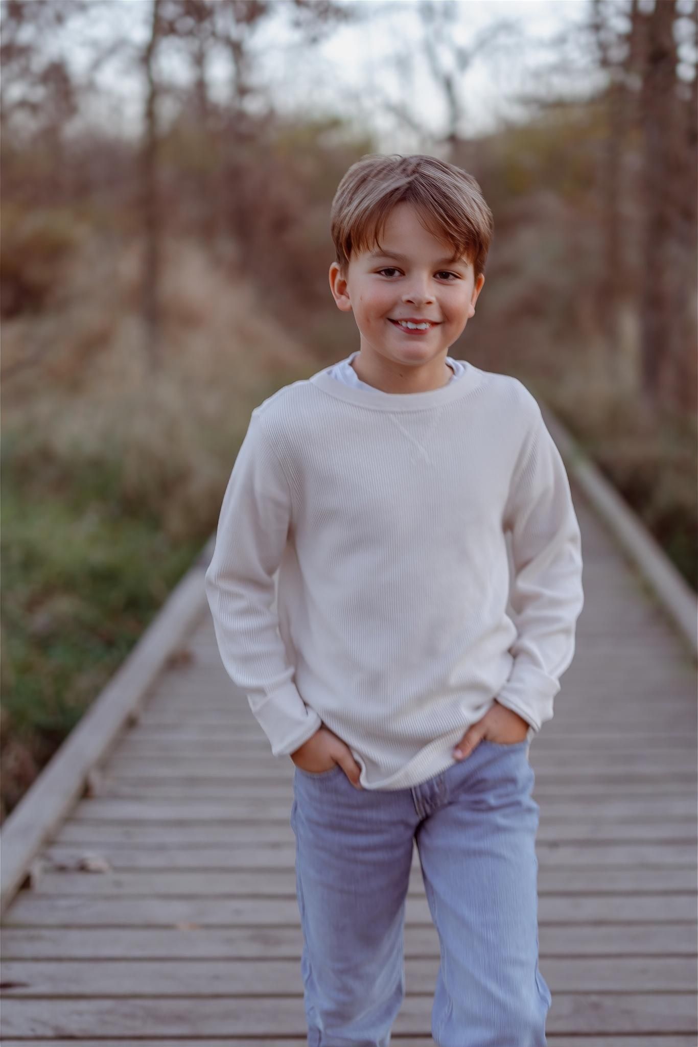 Smiling boy in white sweatshirt and jeans standing on a wooden boardwalk path in an autumn forest.