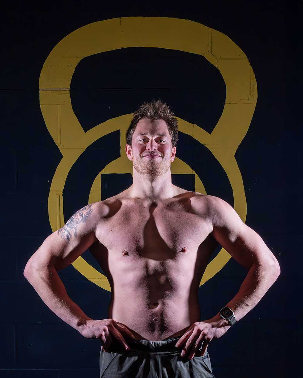 Editorial fitnessportret van een gespierde mannelijke atleet in frontale pose, met nadruk op borst, armen en core.