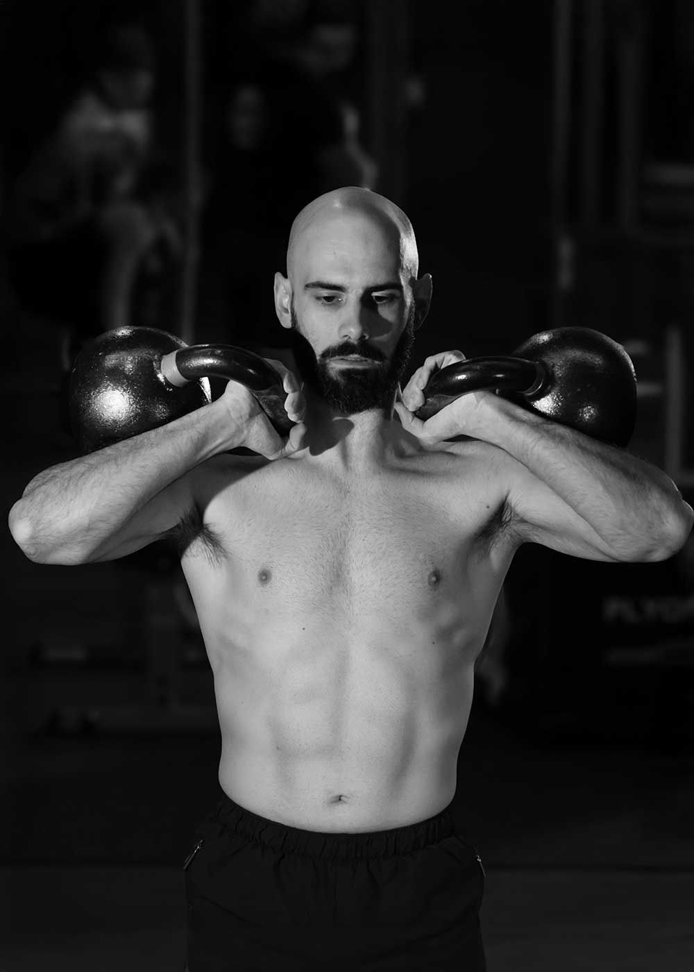 Sfeervol zwart-wit portret van een mannelijke atleet met kettlebells op schouderhoogte, met nadruk op kracht, balans en gecontroleerde power