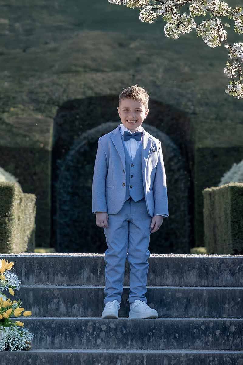 Communieportret van een jonge jongen die op stenen trappen staat, met een klassieke outfit en een rustige buitenomgeving.