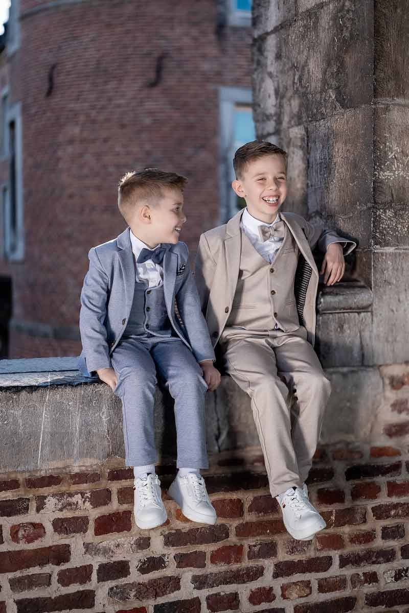 Communiefotografie van twee broers die op een bakstenen muur zitten, met elegante outfits en een ontspannen familieband.