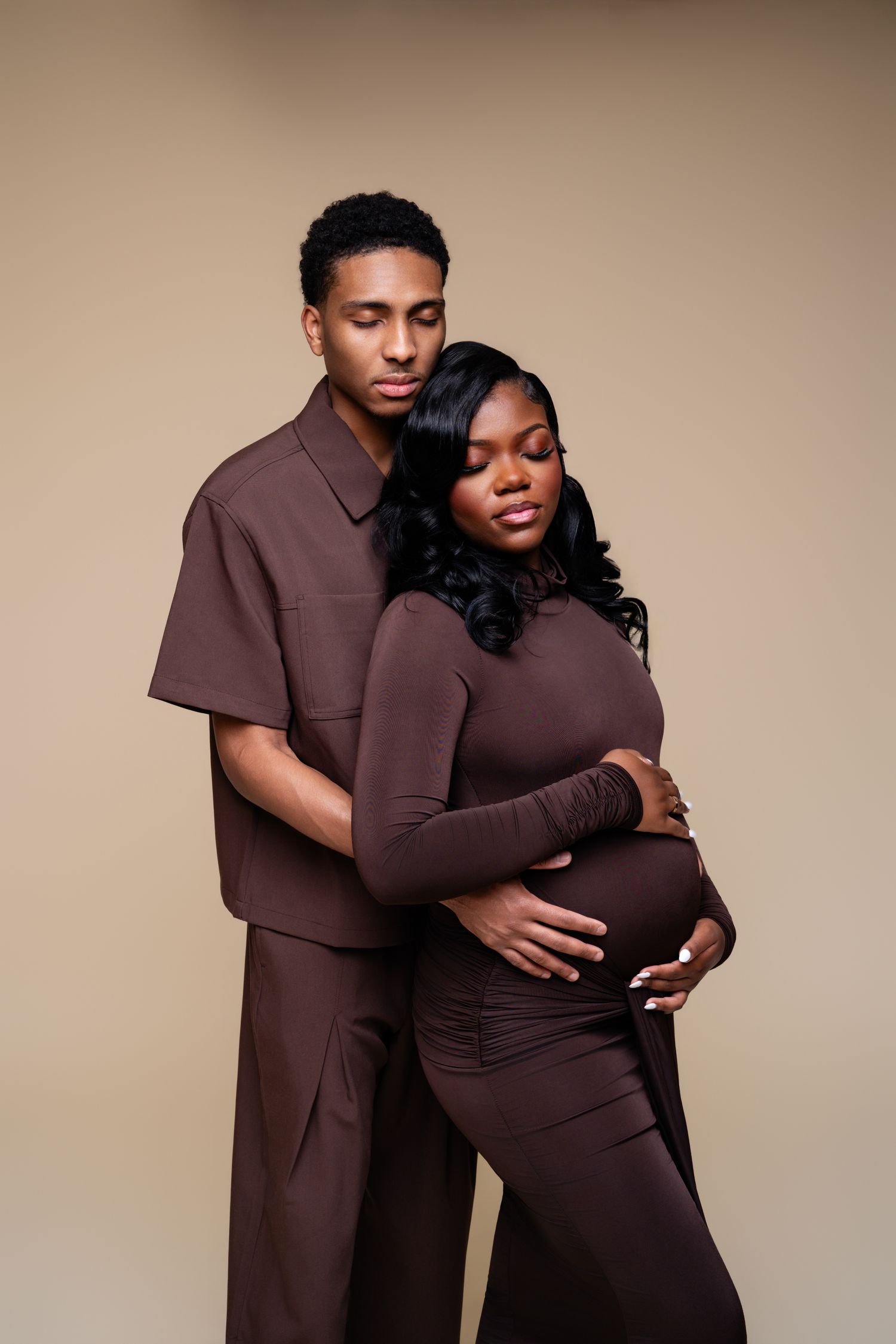 A pregnant Black woman in brown outfit embraced by her partner, both wearing matching brown ensembles on beige background.