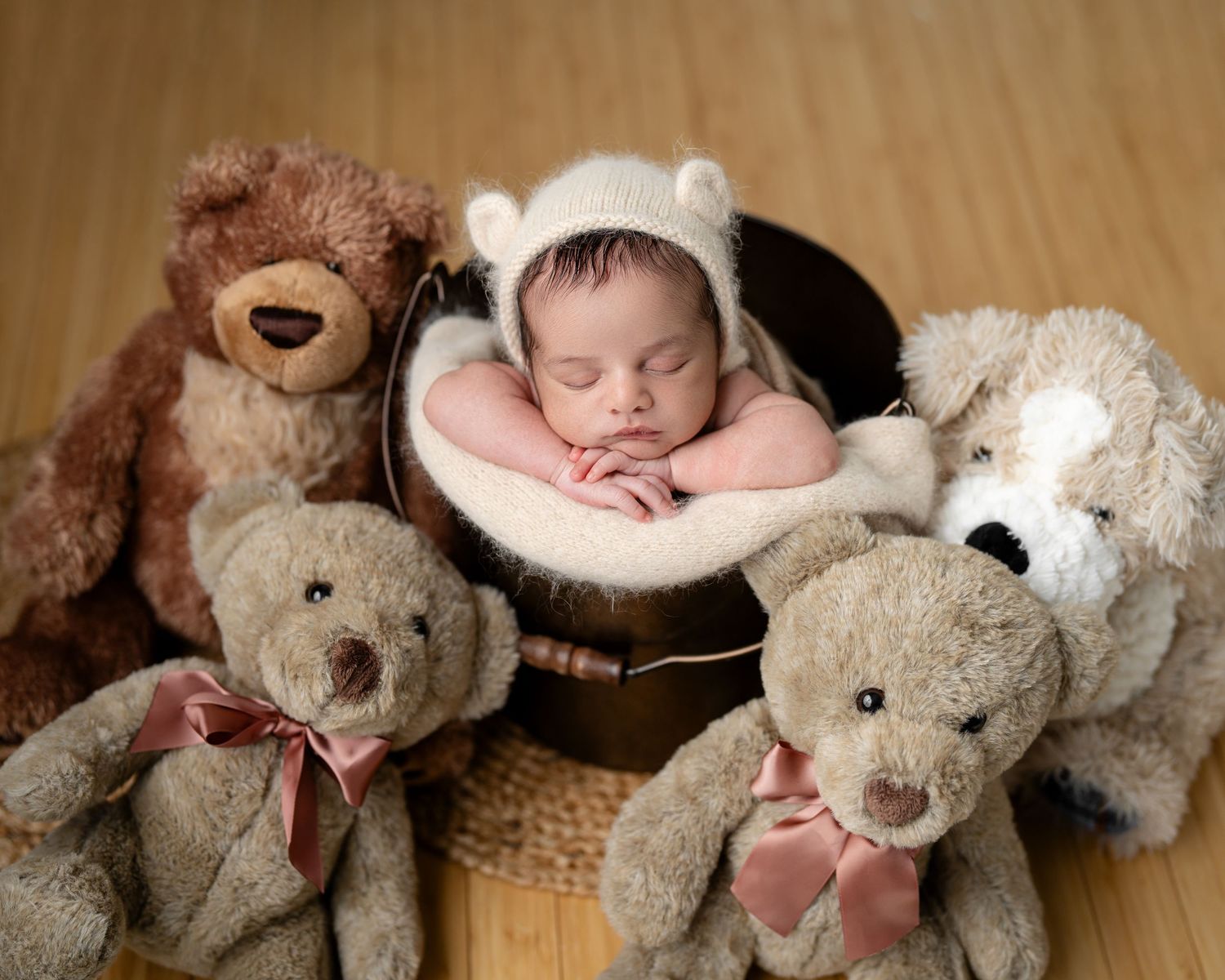 Sleeping newborn baby wearing a bear bonnet rests in a bucket surrounded by four soft teddy bears on a wood floor.