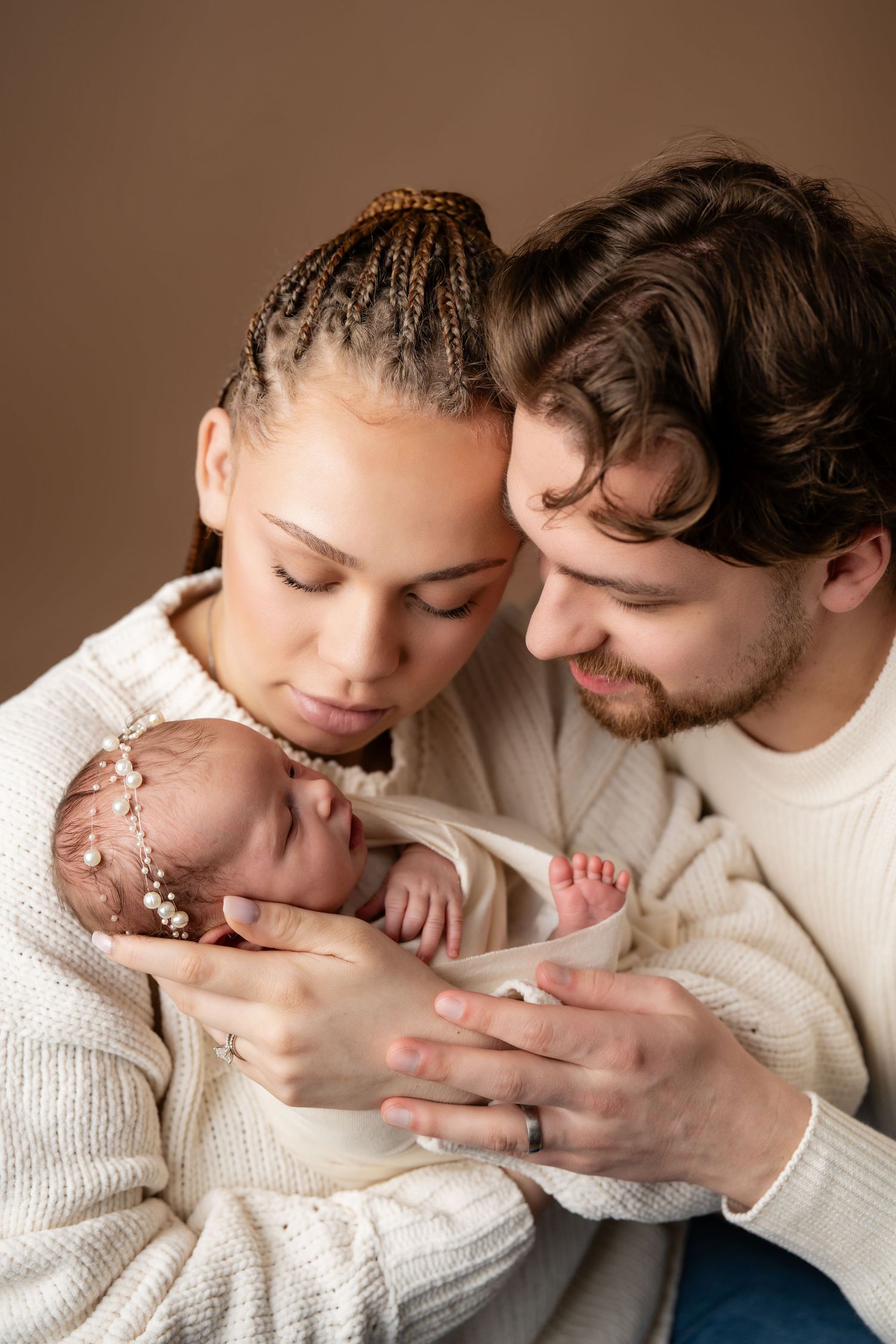 A couple tenderly holds their newborn baby, dressed in white knit sweaters, against a warm brown background.