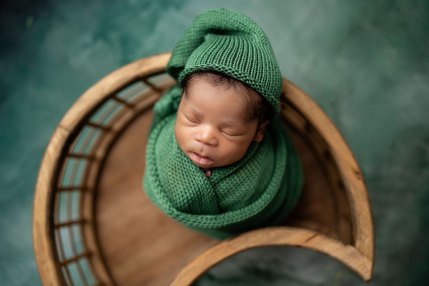 Newborn baby swaddled in green knit wrap and hat, sleeping peacefully in a wooden bowl prop.