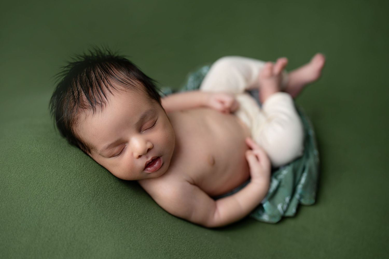 Newborn baby sleeping peacefully on green backdrop, wearing white pants, with dark hair and a subtle smile.