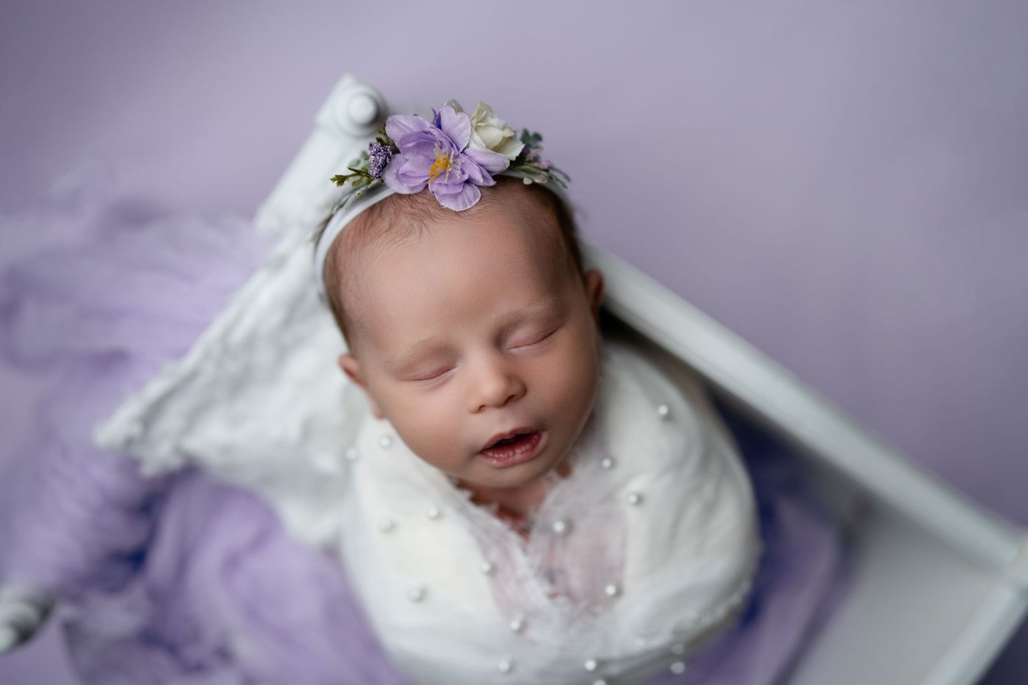Sleeping newborn baby wrapped in white swaddle with purple floral headband in a white wooden box.