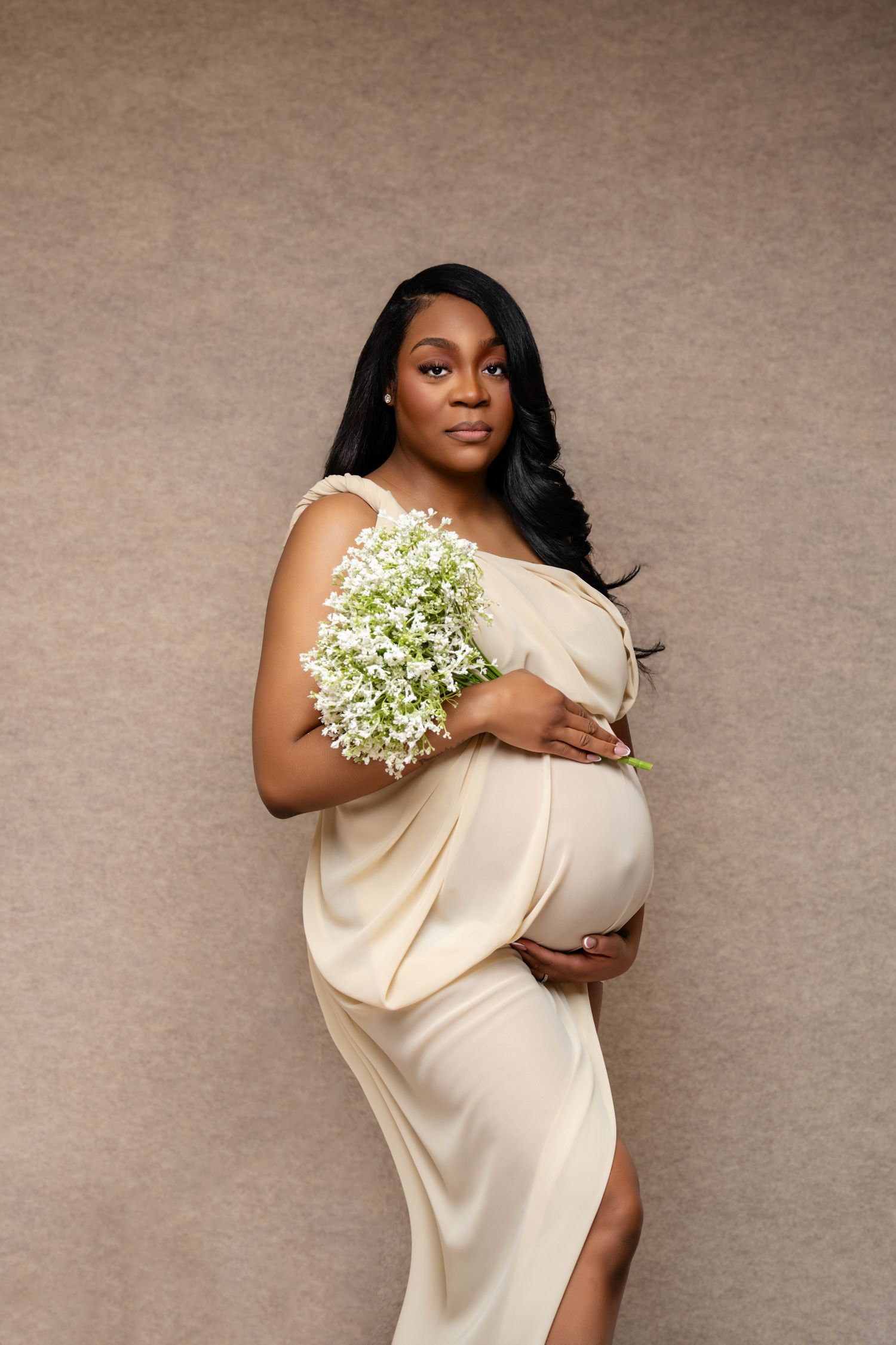 Pregnant Black woman in elegant cream draped gown holding white baby's breath flowers, posing gracefully against a neutral background.