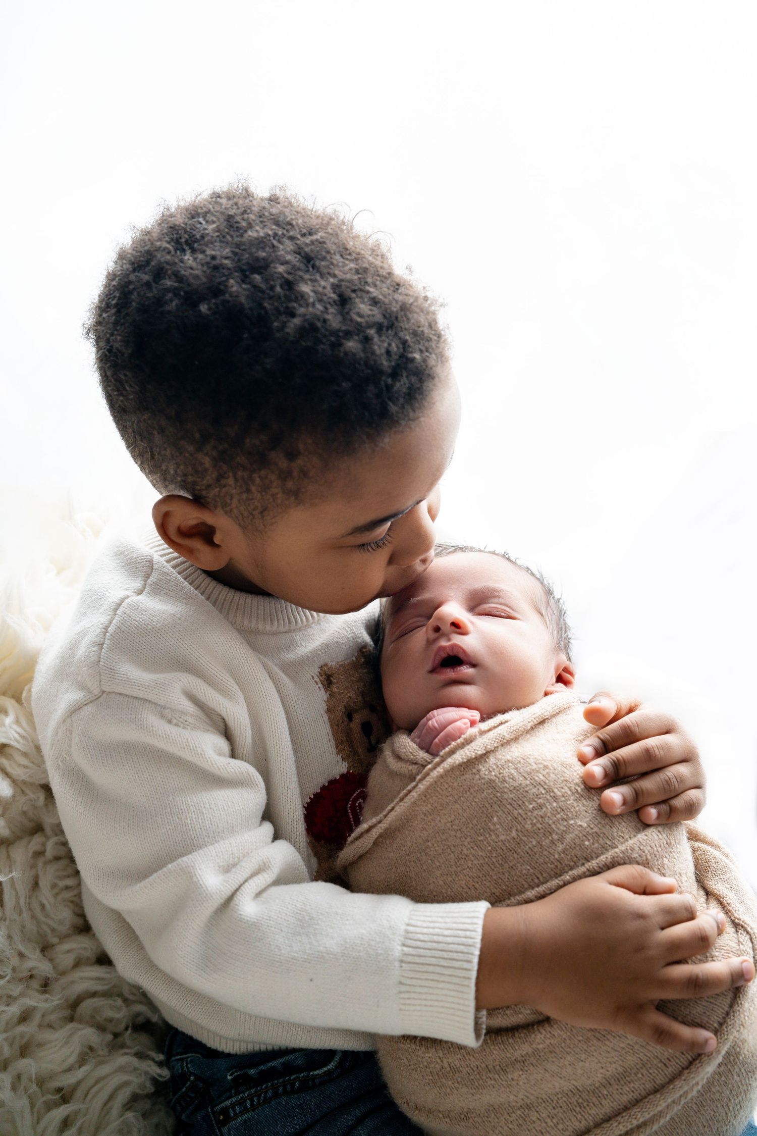 Young boy tenderly kissing newborn baby sibling while gently cradling them against white background.