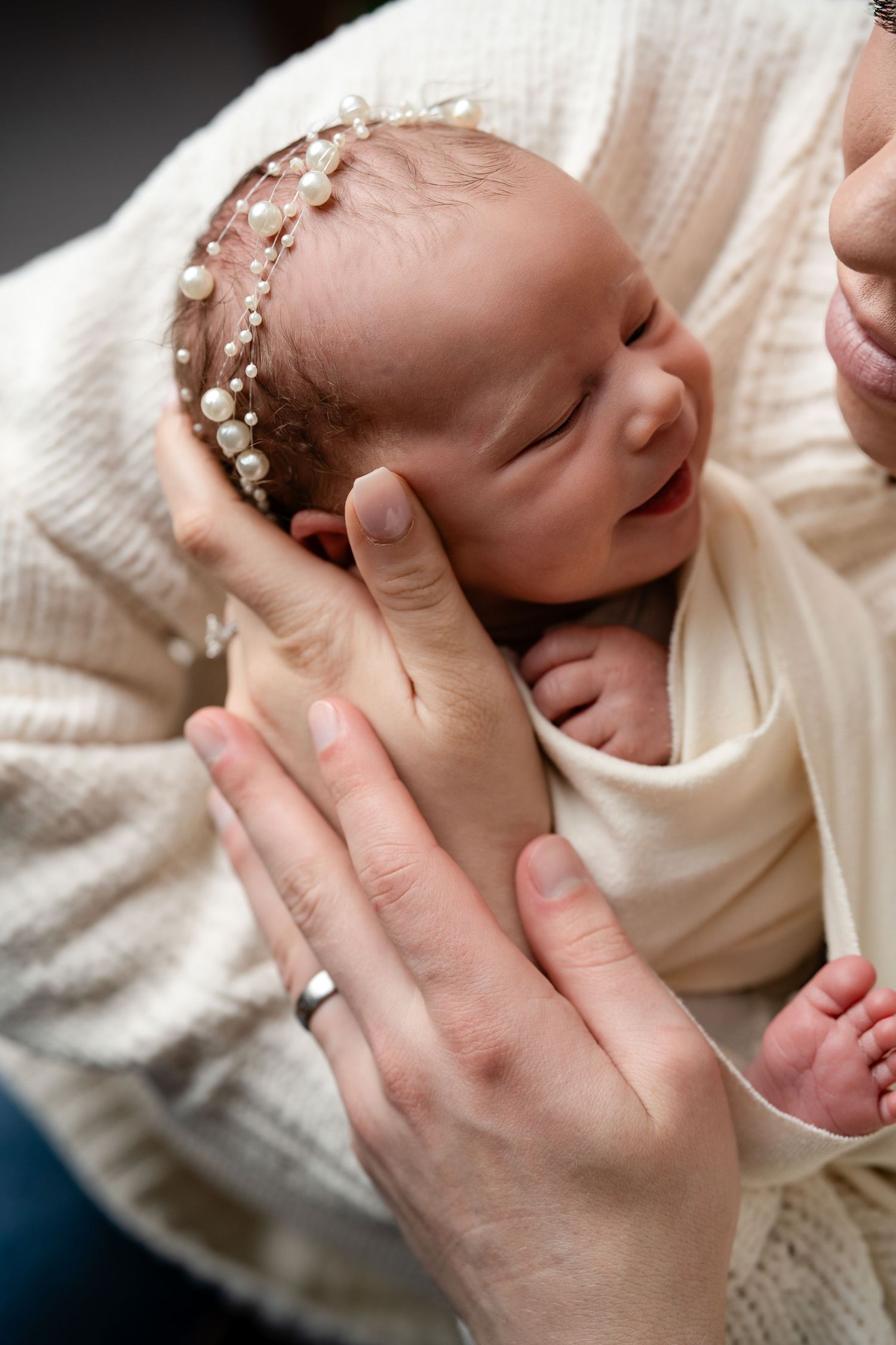 Newborn baby girl wearing pearl headband wrapped in cream swaddle, cradled in parent's gentle hands.
